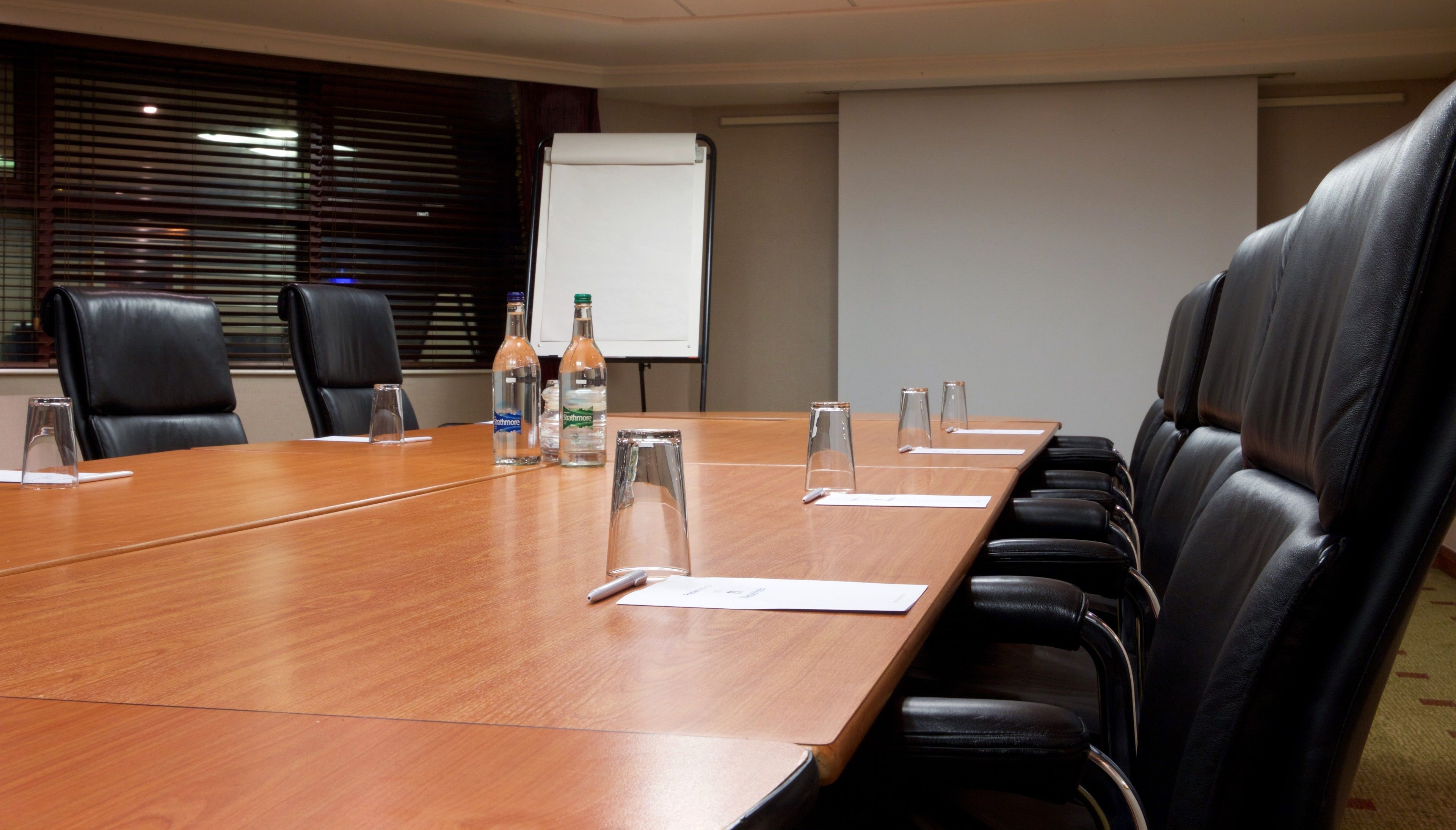Un salón de conferencias con una larga mesa de madera, sillas de cuero negro, botellas de agua y una pizarra.