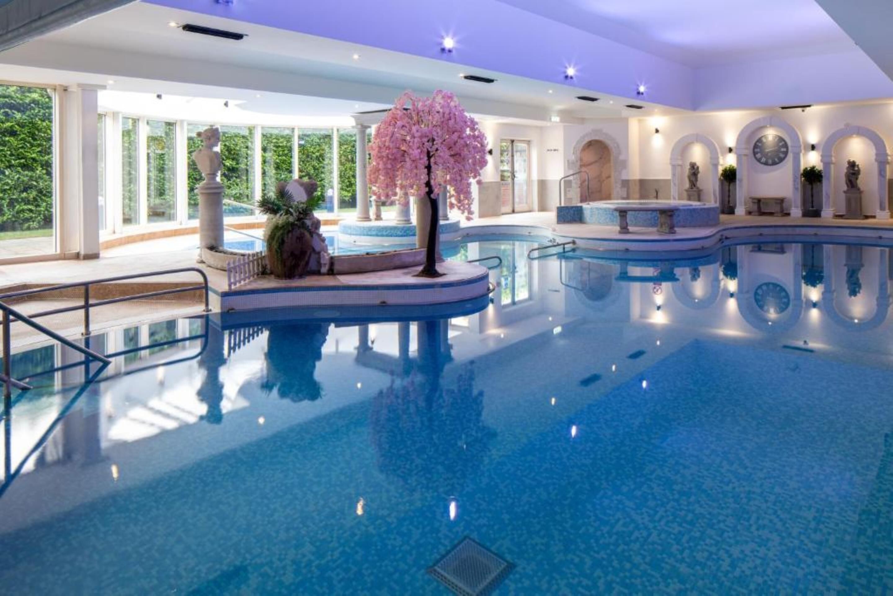 A large indoor swimming pool with a central island, clear glass railings, and a view of greenery outside.