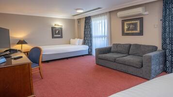 A hotel room with a bed, a grey sofa, a desk with a lamp, and two framed pictures on the walls.