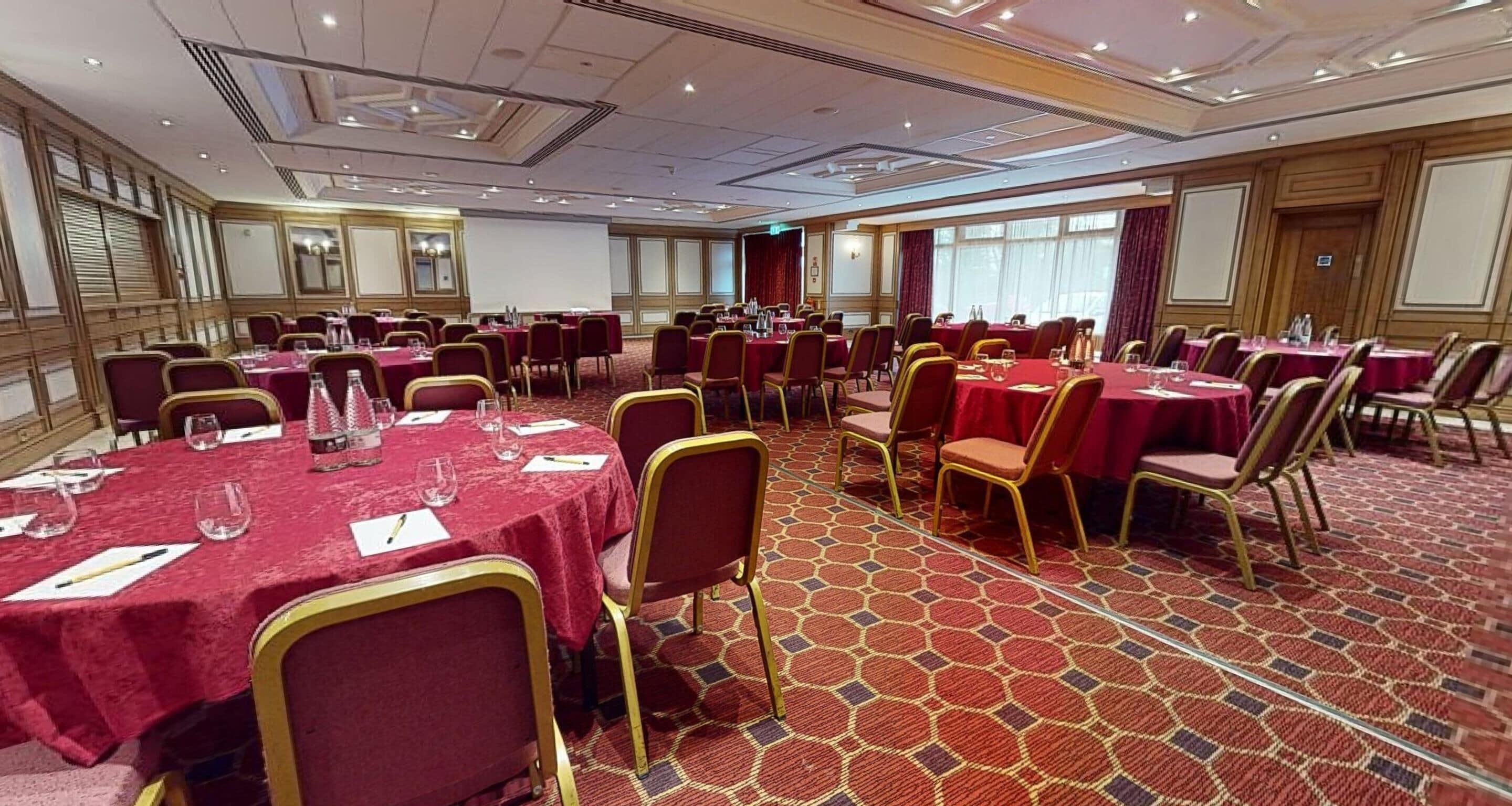 A conference room with multiple tables and chairs, carpeted floor, and large windows.