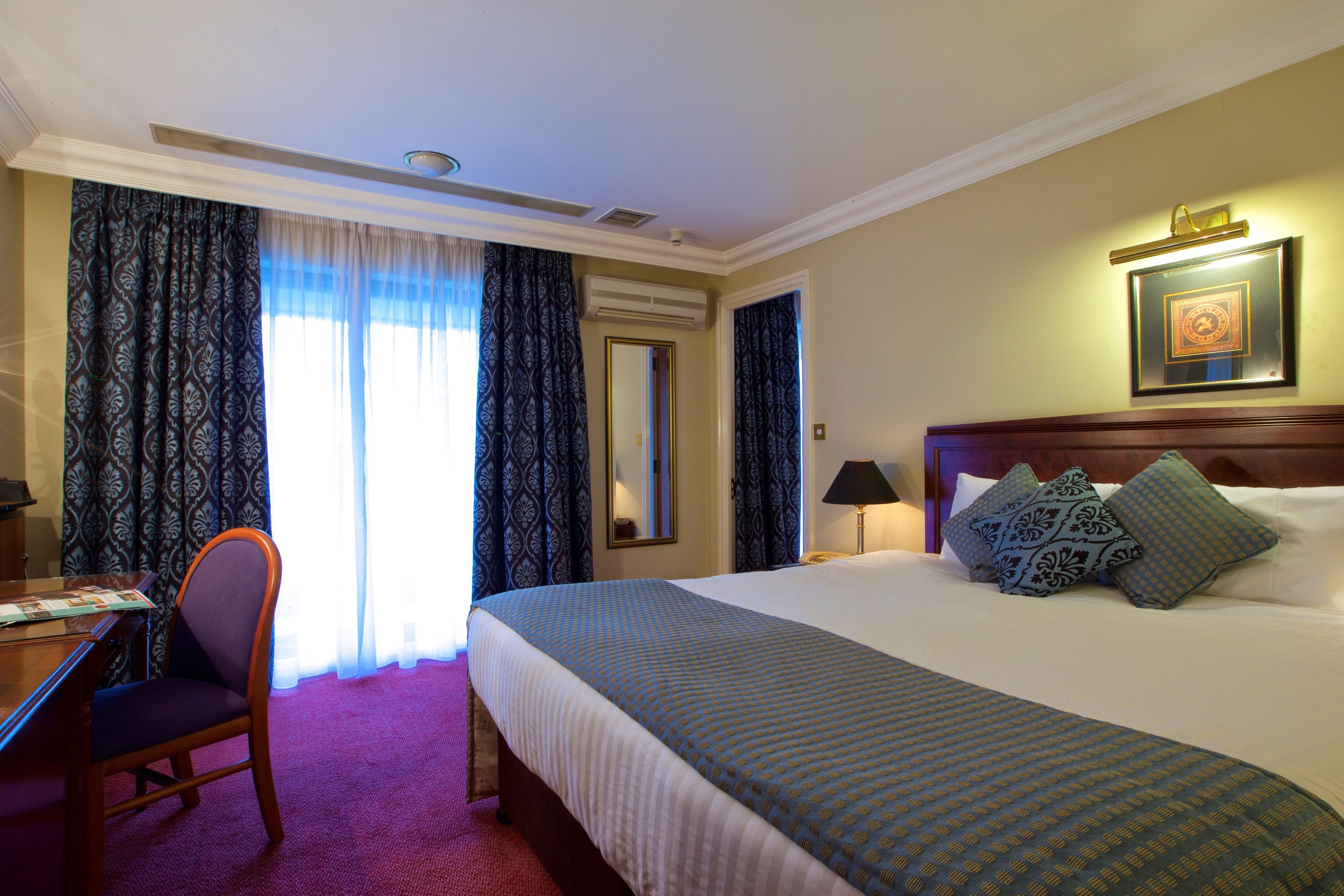 Habitación de hotel con una cama grande, un escritorio y una silla. Cuenta con una ventana con cortinas, un marco de cuadros en la pared y una mesita de noche con una lámpara.