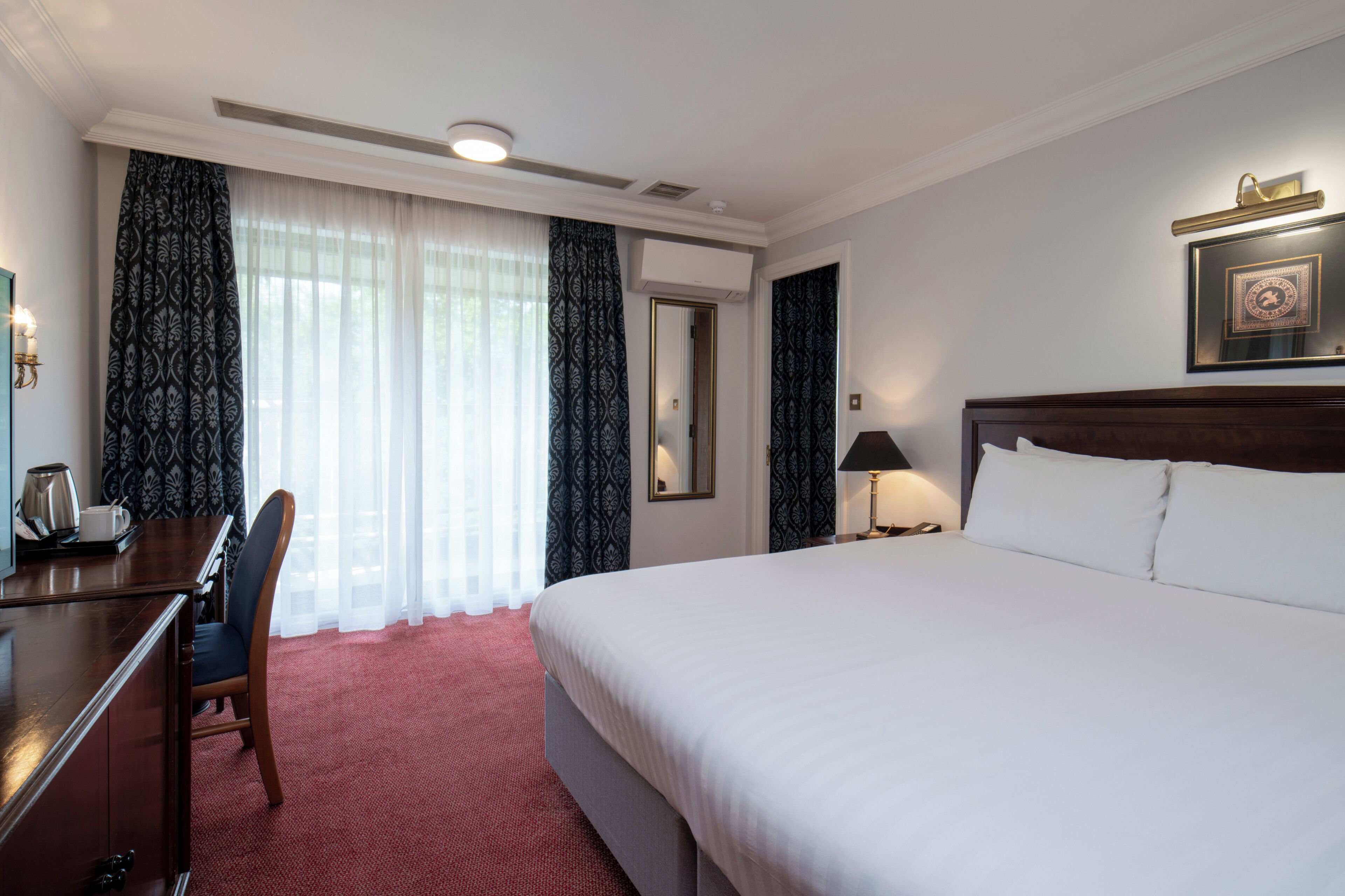 A hotel room with a large bed, a desk with a chair, a bedside lamp, and a framed picture on the wall.
