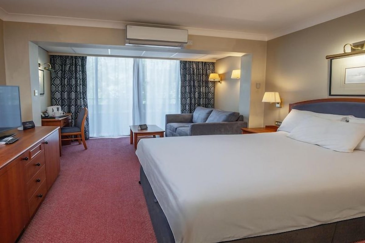 A hotel room with a bed, a desk with a TV, a chair, a small table, and a window with curtains.