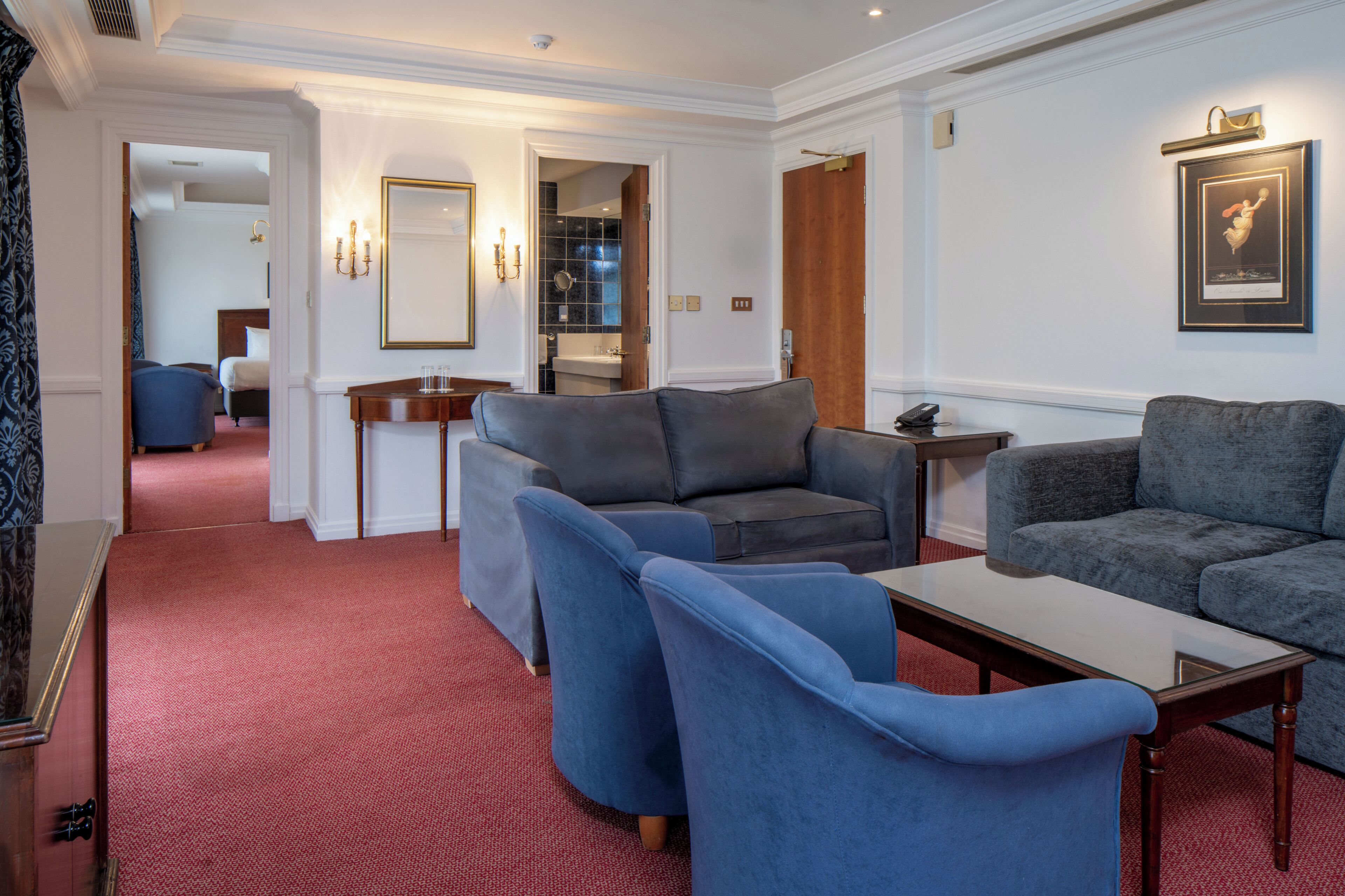 A hotel room with a red carpet, two grey sofas, a small table, and a mirror.