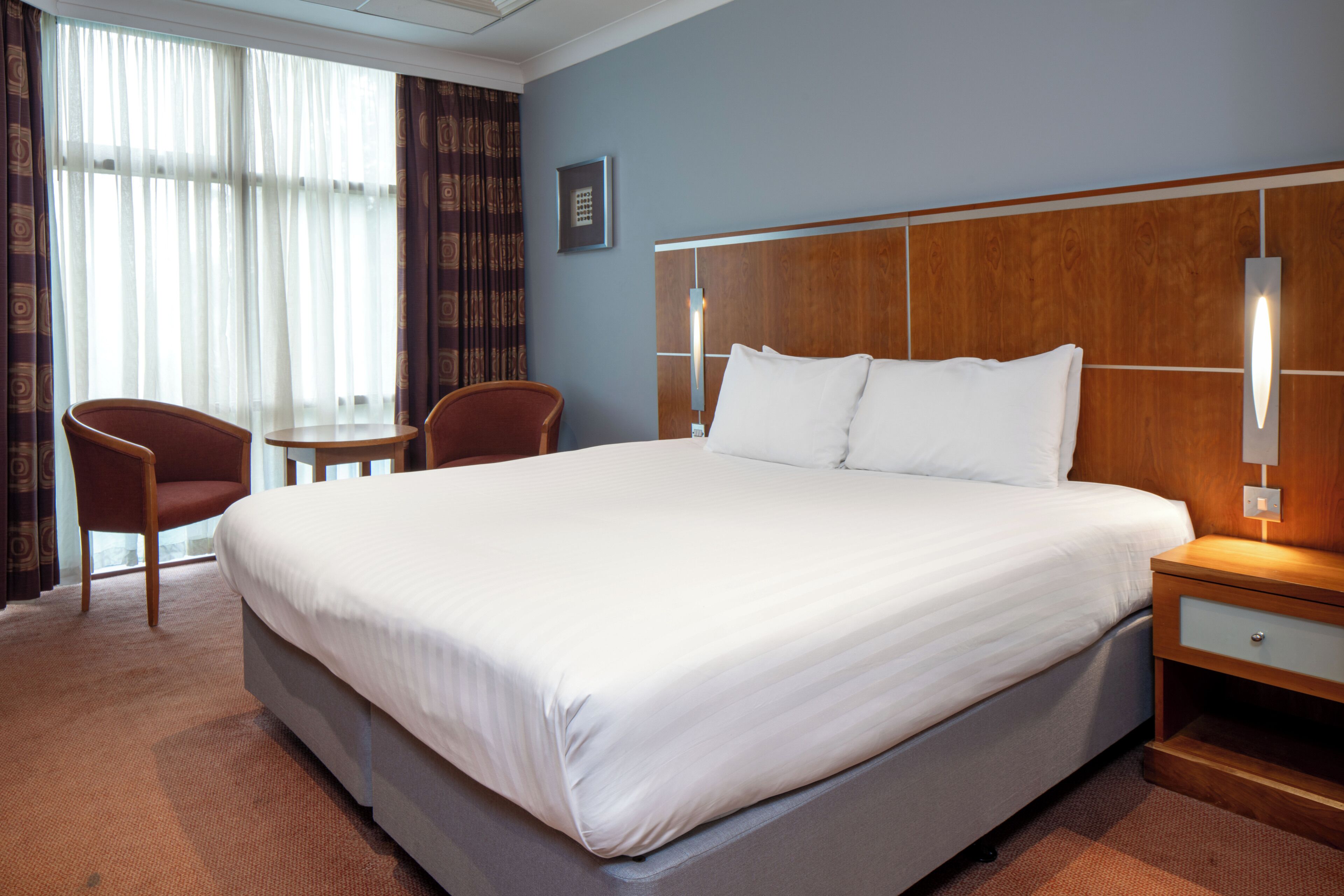A hotel room with a large bed, two chairs, a small table, and a nightstand with a lamp.