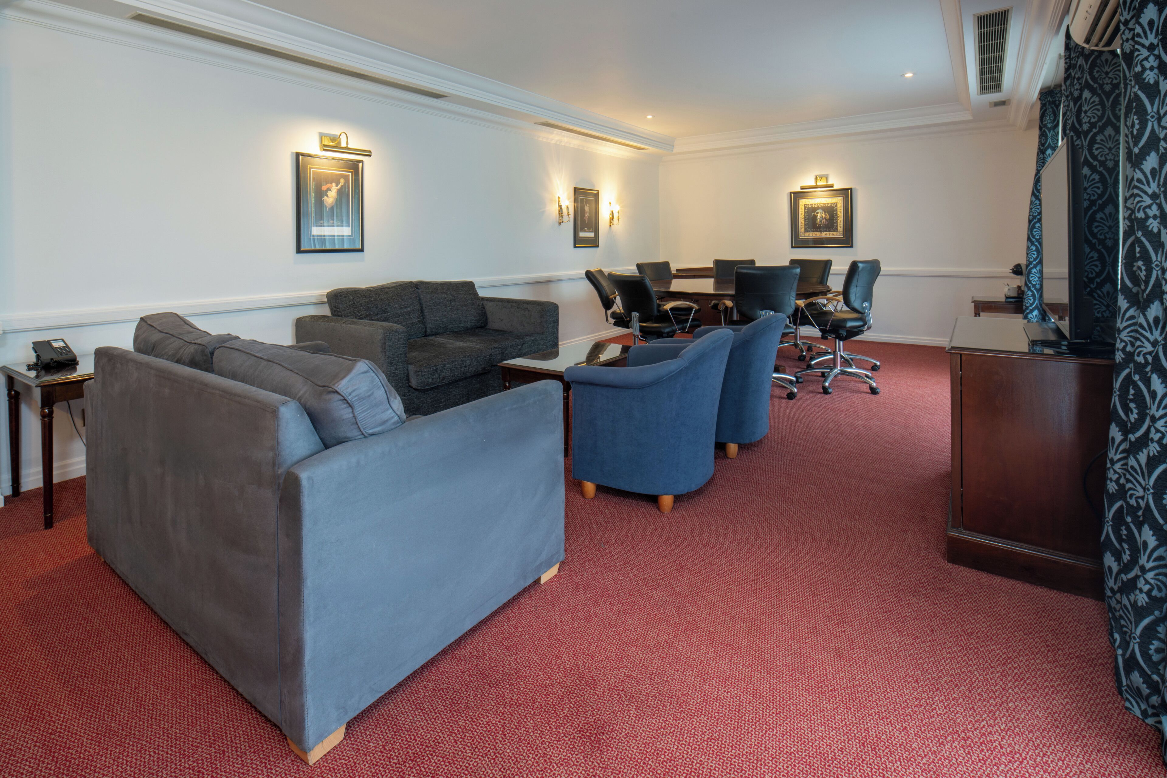 Een hotelkamer met een rode vloerkleed, grijze en blauwe banken, een houten bijzettafeltje en een vergadertafel met stoelen.
