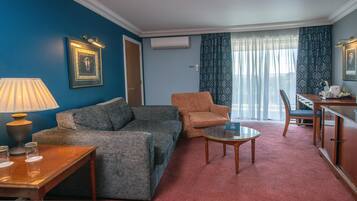 A hotel room with a grey sofa, a small round table, a wooden cabinet, a lamp, and a framed picture on the wall.