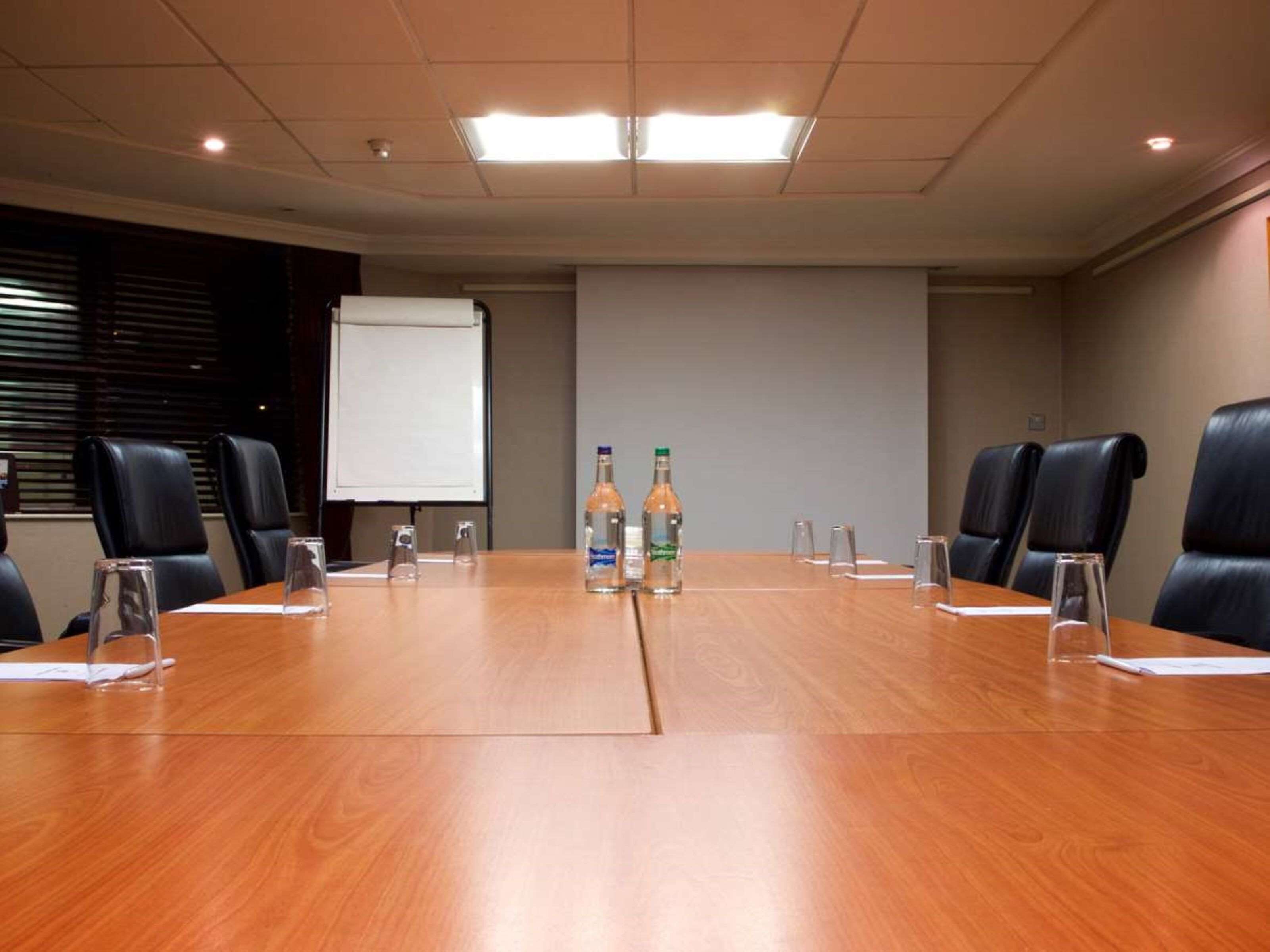 Une salle de conférence avec une longue table en bois, des fauteuils en cuir noir, un écran de projection et deux bouteilles d’eau.