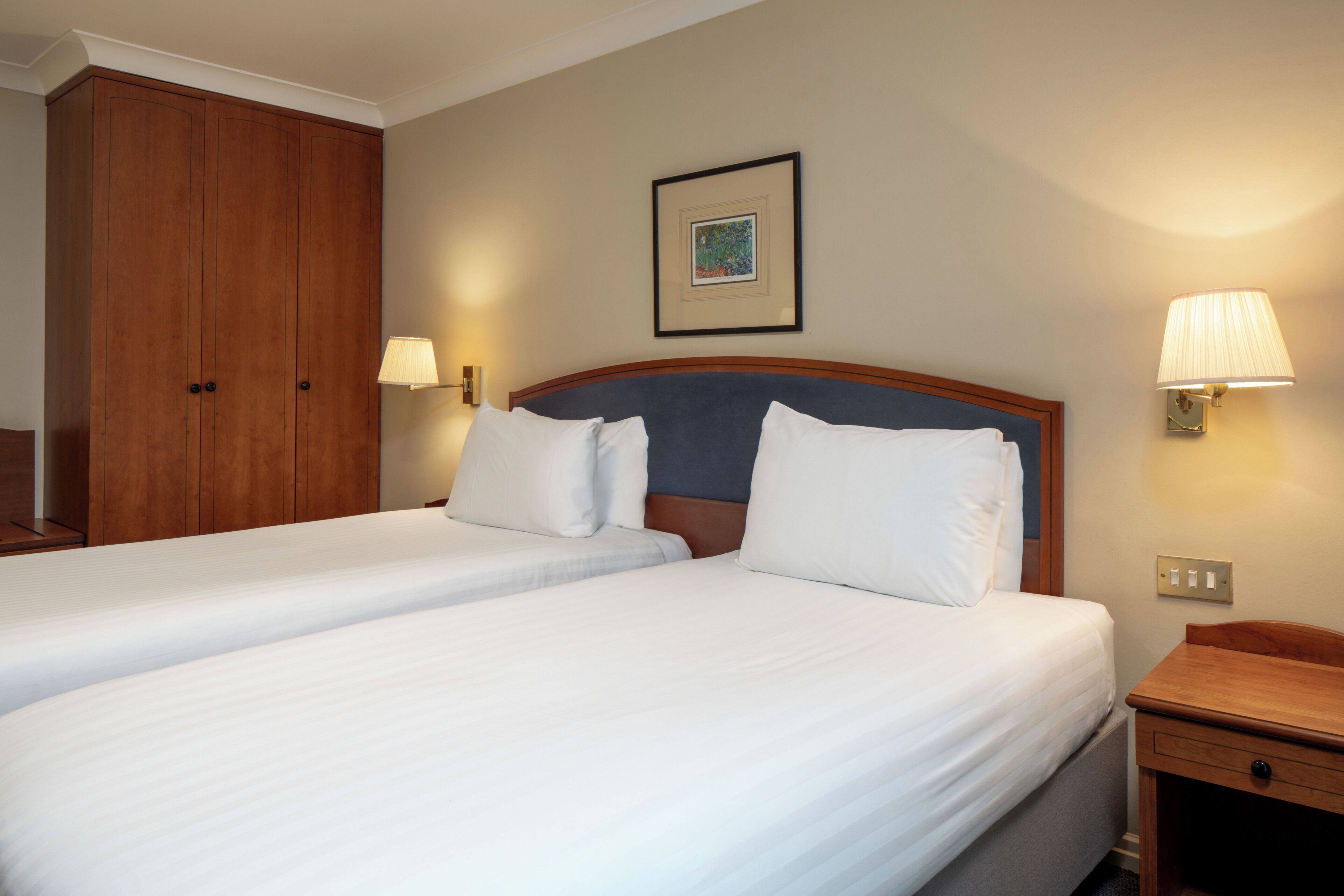 Ein Hotelzimmer mit zwei Betten, einem Holzschrank, einem gerahmten Bild an der Wand und Nachttischlampen.
