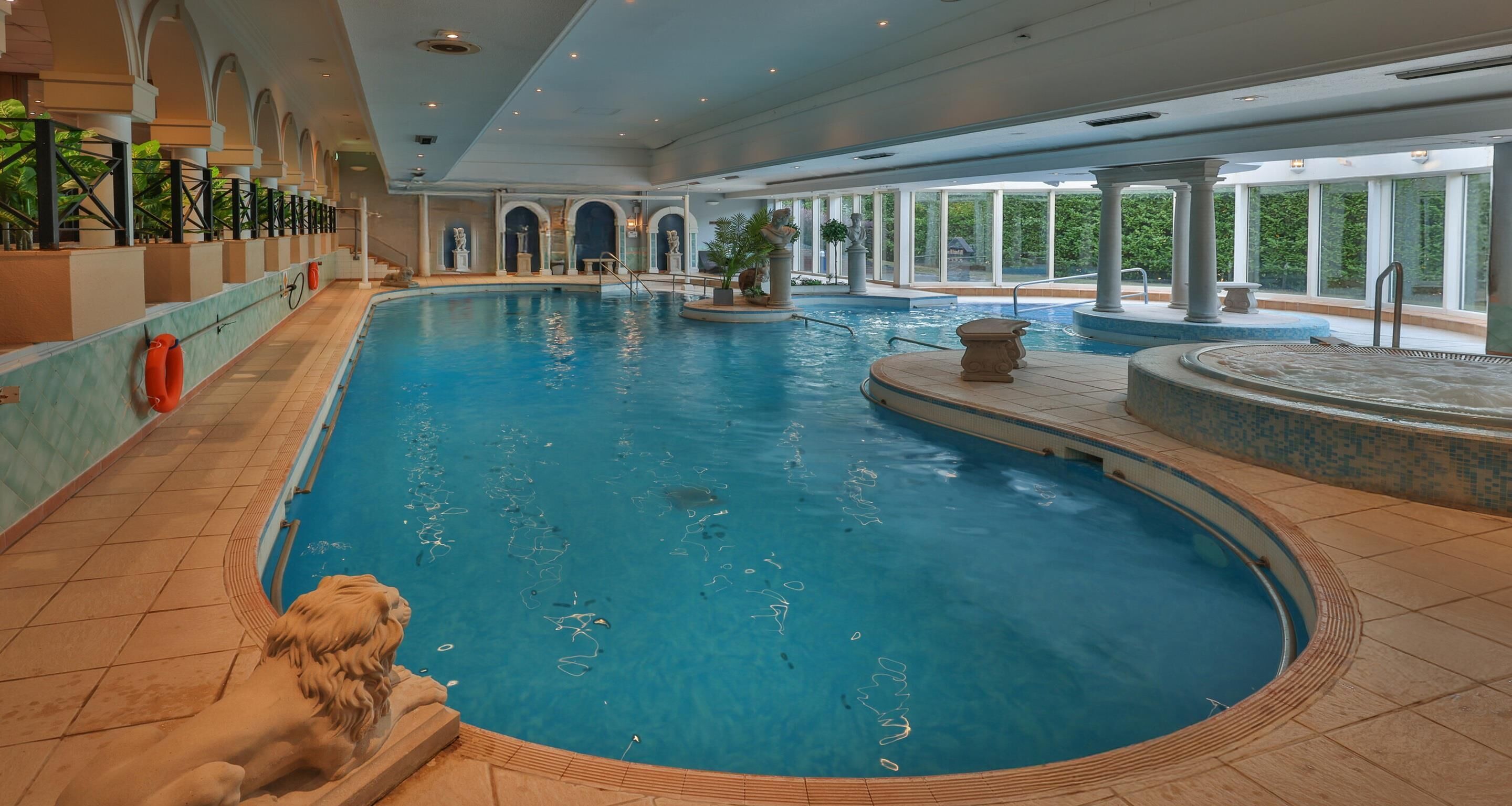 A large indoor swimming pool with clear blue water, surrounded by a tiled floor and a stone lion statue.