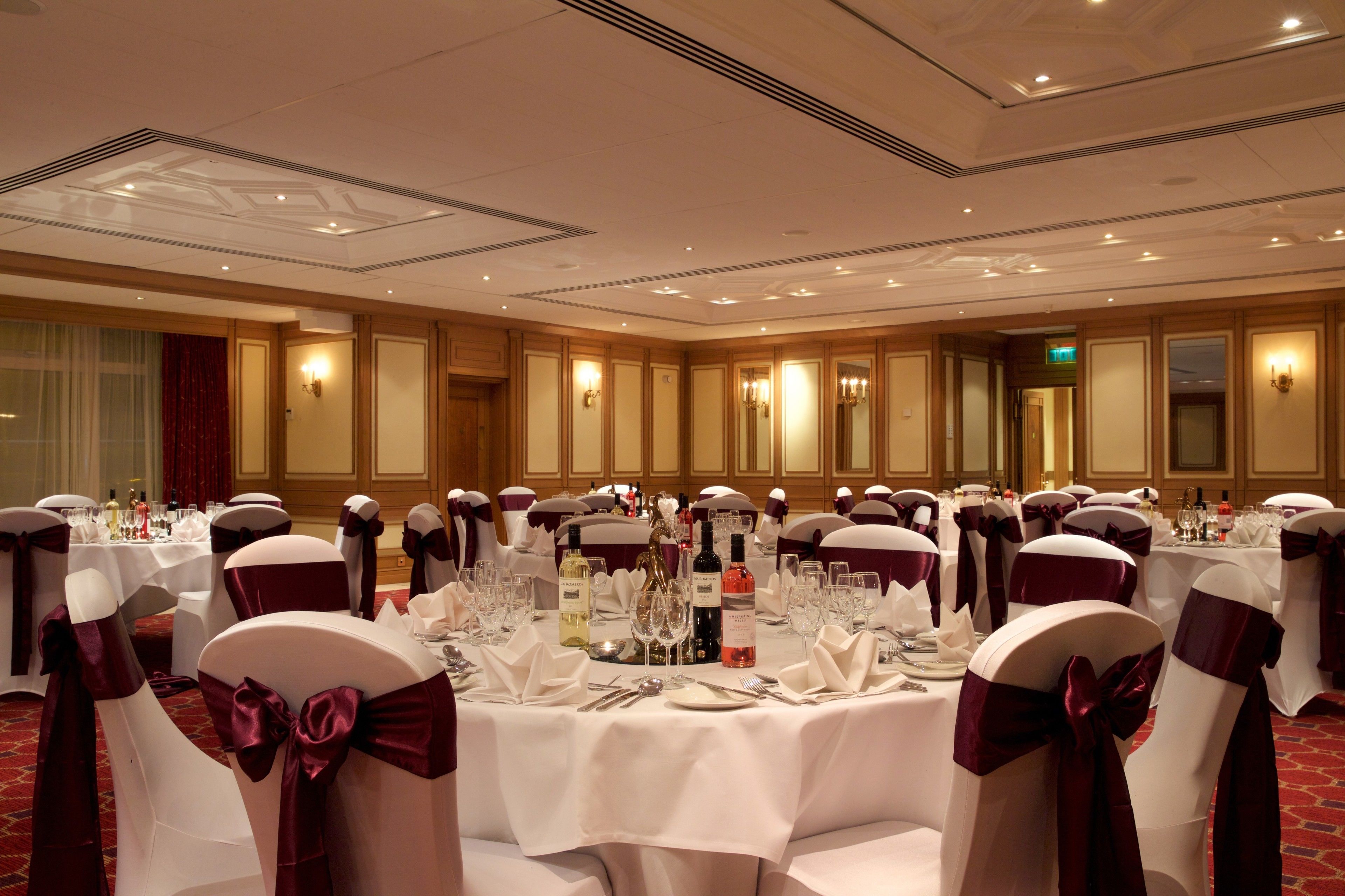 Une salle de banquet avec des tables rondes dressées pour un événement formel, avec des nappes blanches et des rubans rouges sur les chaises.