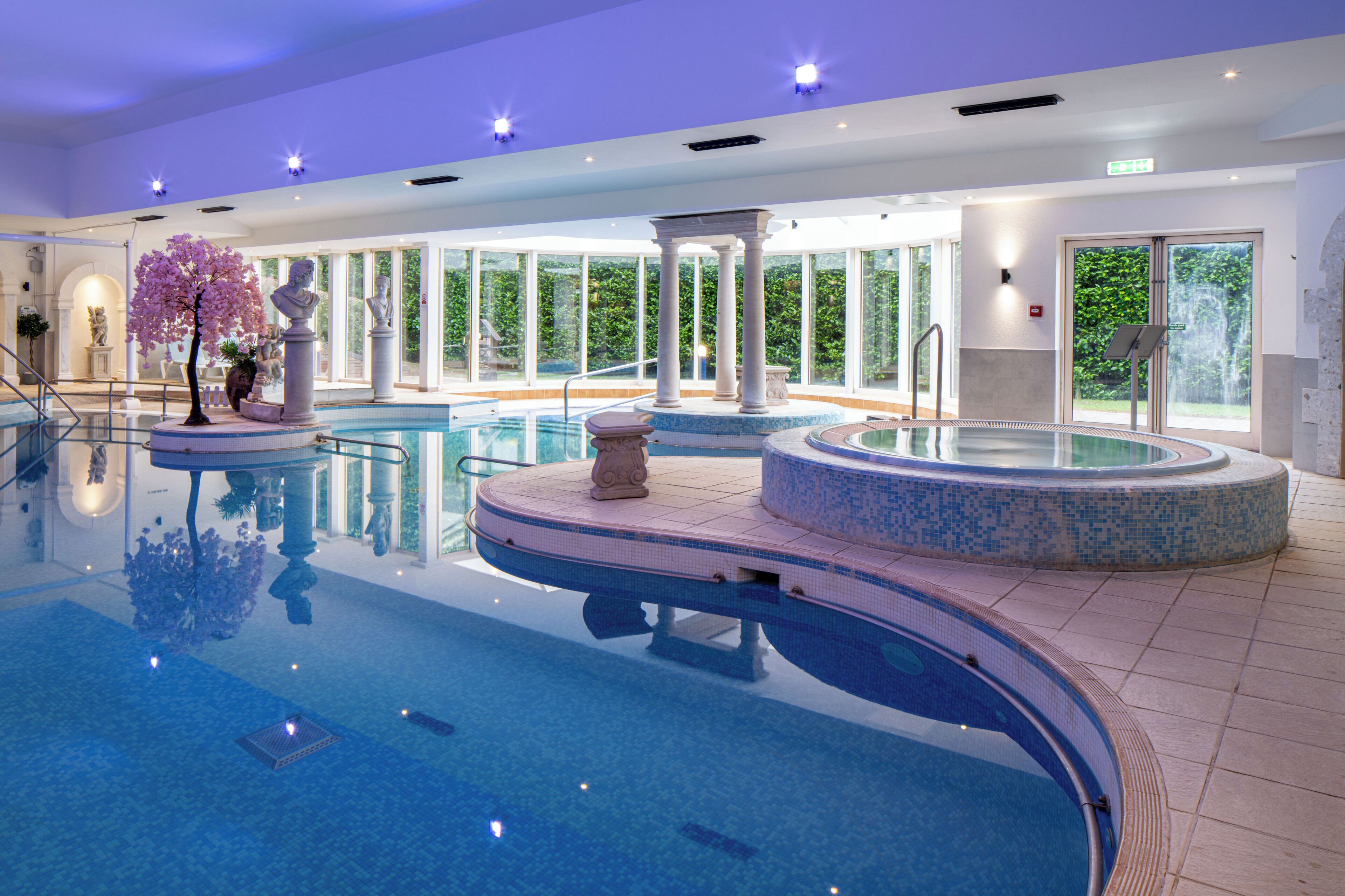 A large indoor swimming pool with clear blue water, surrounded by a tiled deck and a glass enclosure.