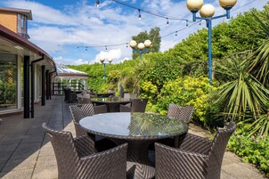 An outdoor patio area with round tables and wicker chairs, surrounded by lush greenery and a clear sky.