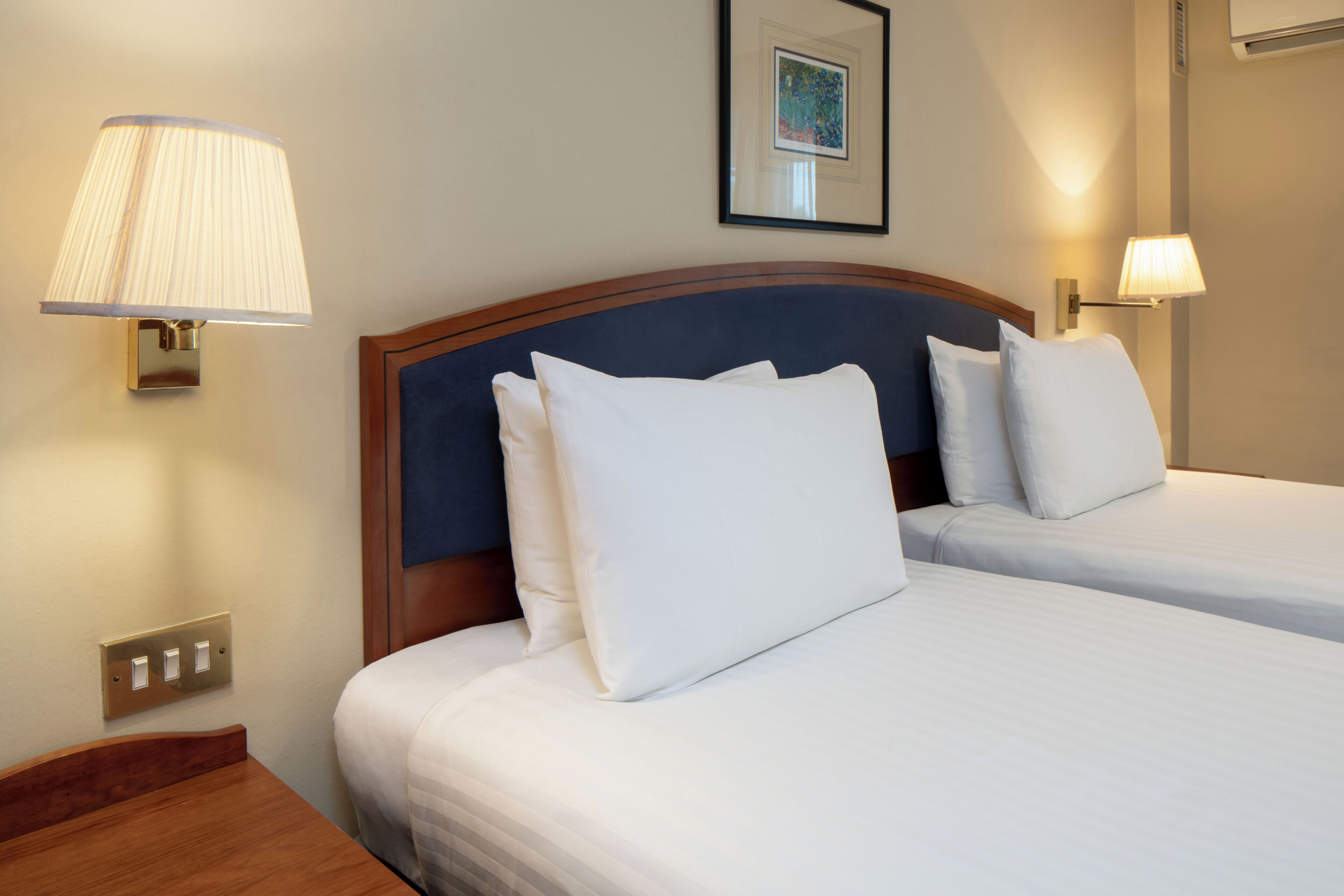 A hotel room with two beds, each with white linens and pillows, a wooden headboard, and wall-mounted lamps.