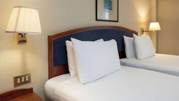 A hotel room with two beds, each with white linens and pillows, a wooden headboard, and wall-mounted lamps.