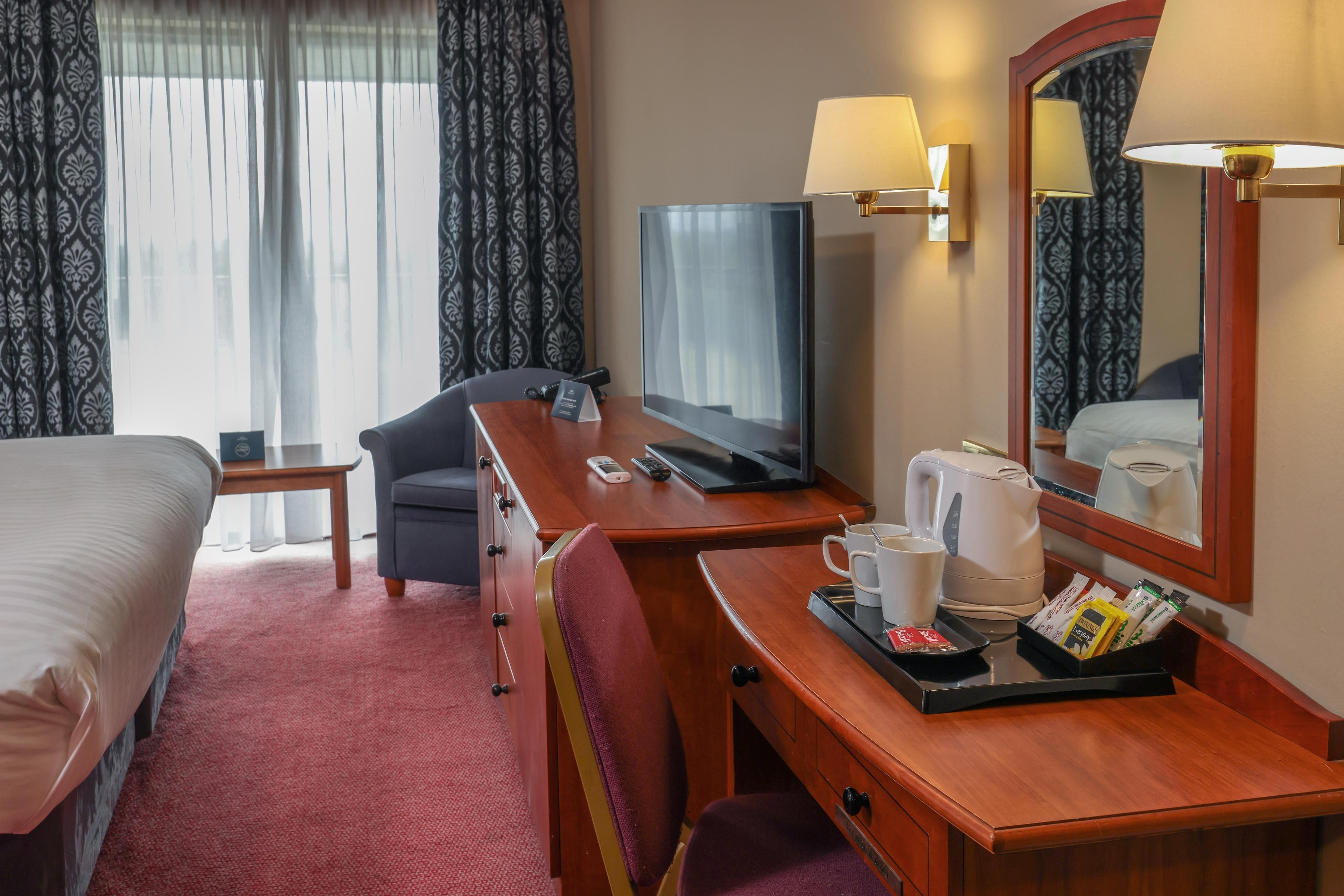 A hotel room with a bed, a desk with a computer, a chair, a television, a coffee maker, and a tray with breakfast items.
