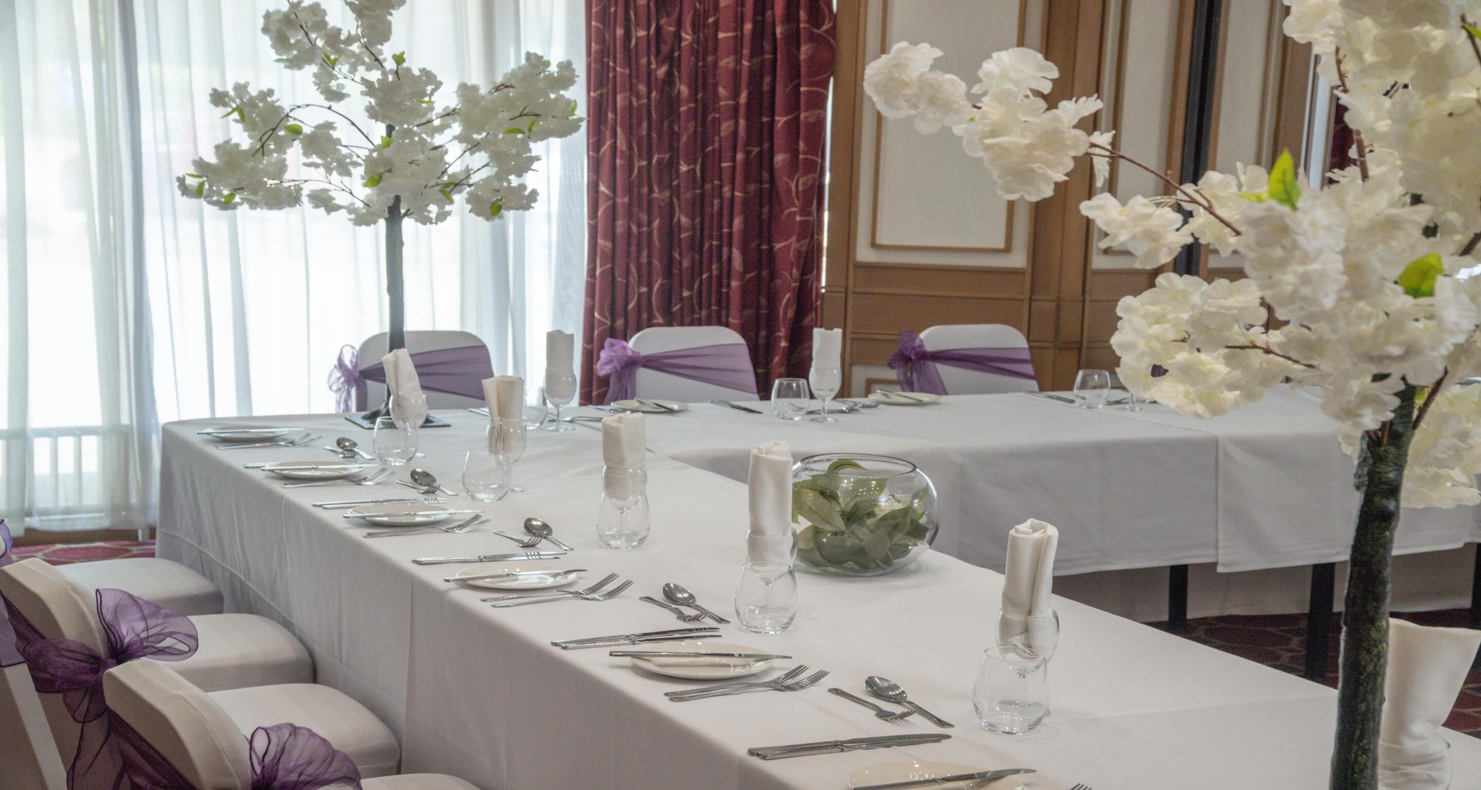 A banquet table set for a formal event with white tablecloths, purple chair sashes, and white floral arrangements.