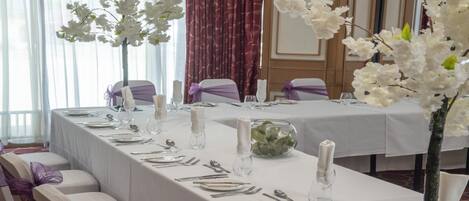 A banquet table set for a formal event with white tablecloths, purple chair sashes, and white floral arrangements.