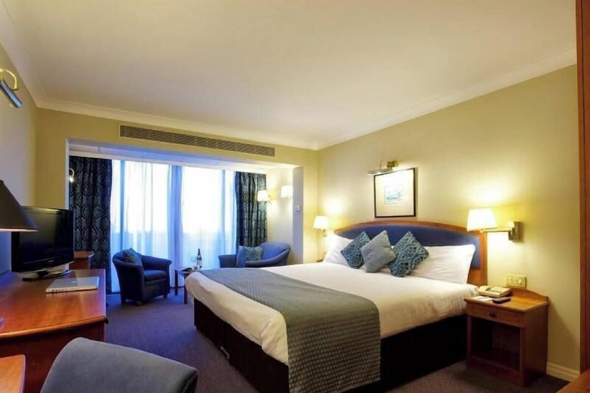 Habitación de hotel con cama, escritorio, silla, televisor y ventana con cortinas.