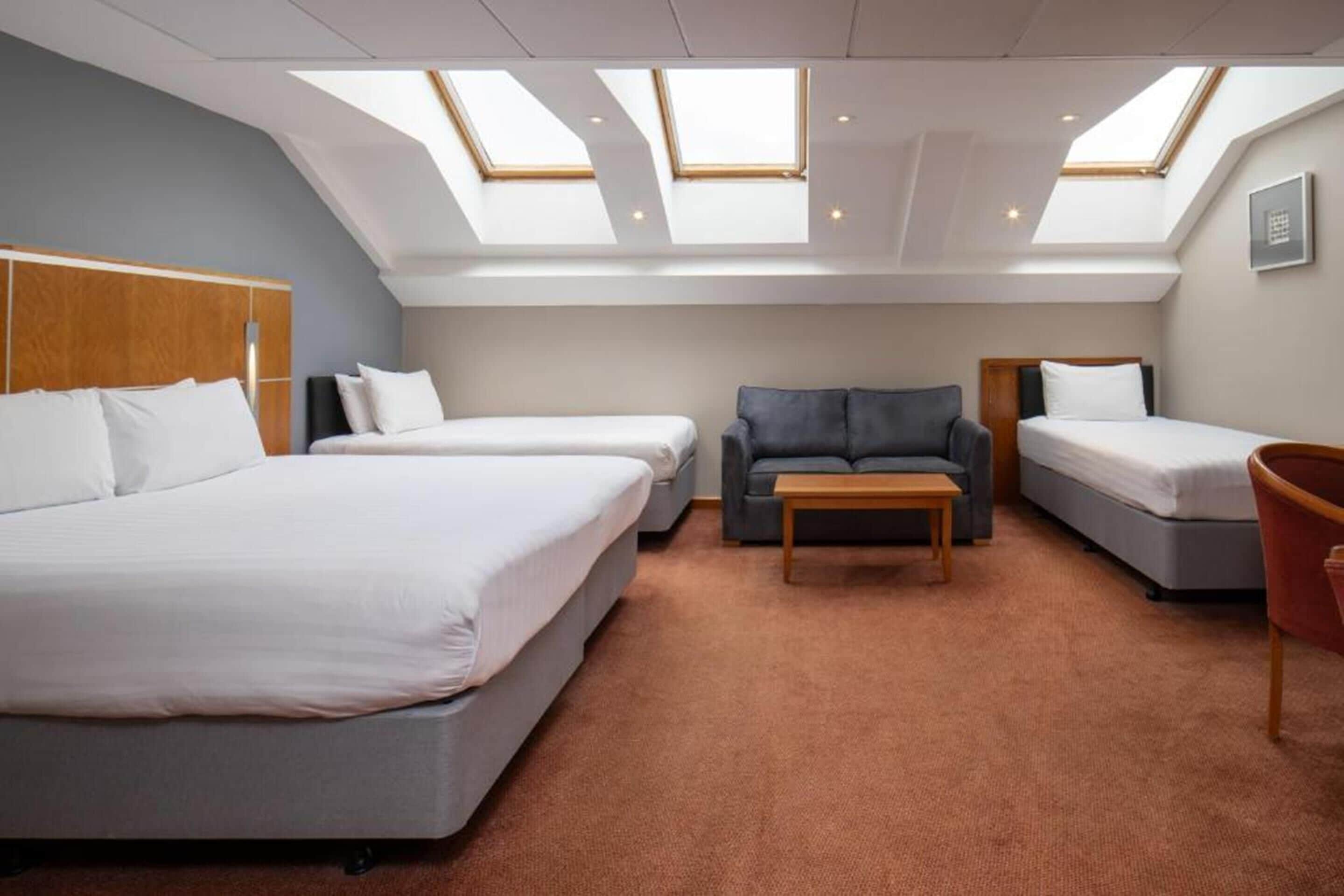 A hotel room with two beds, a sofa, a wooden table, and skylights.