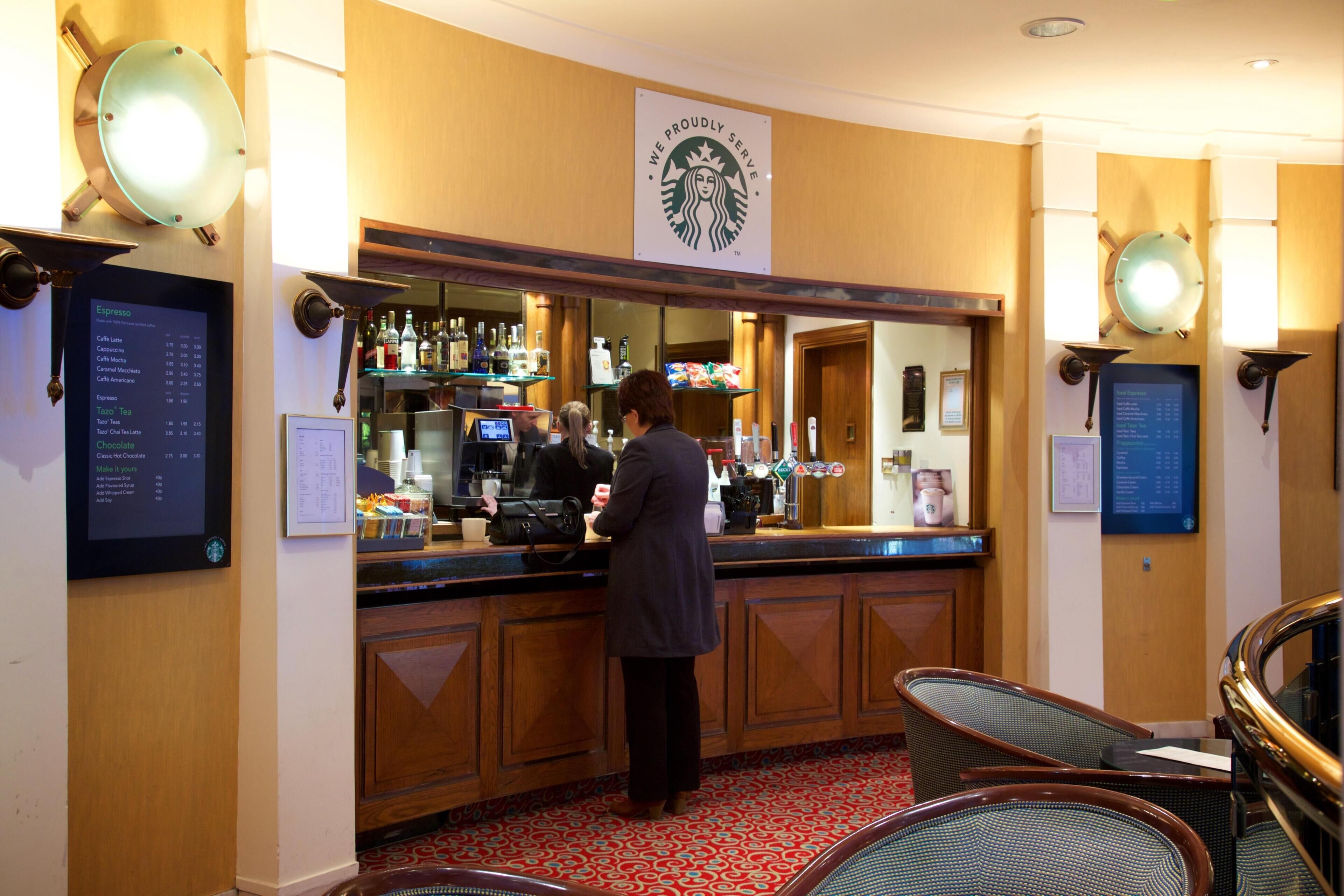 A Starbucks coffee shop with a barista preparing drinks, a menu board, and seating area with chairs.
