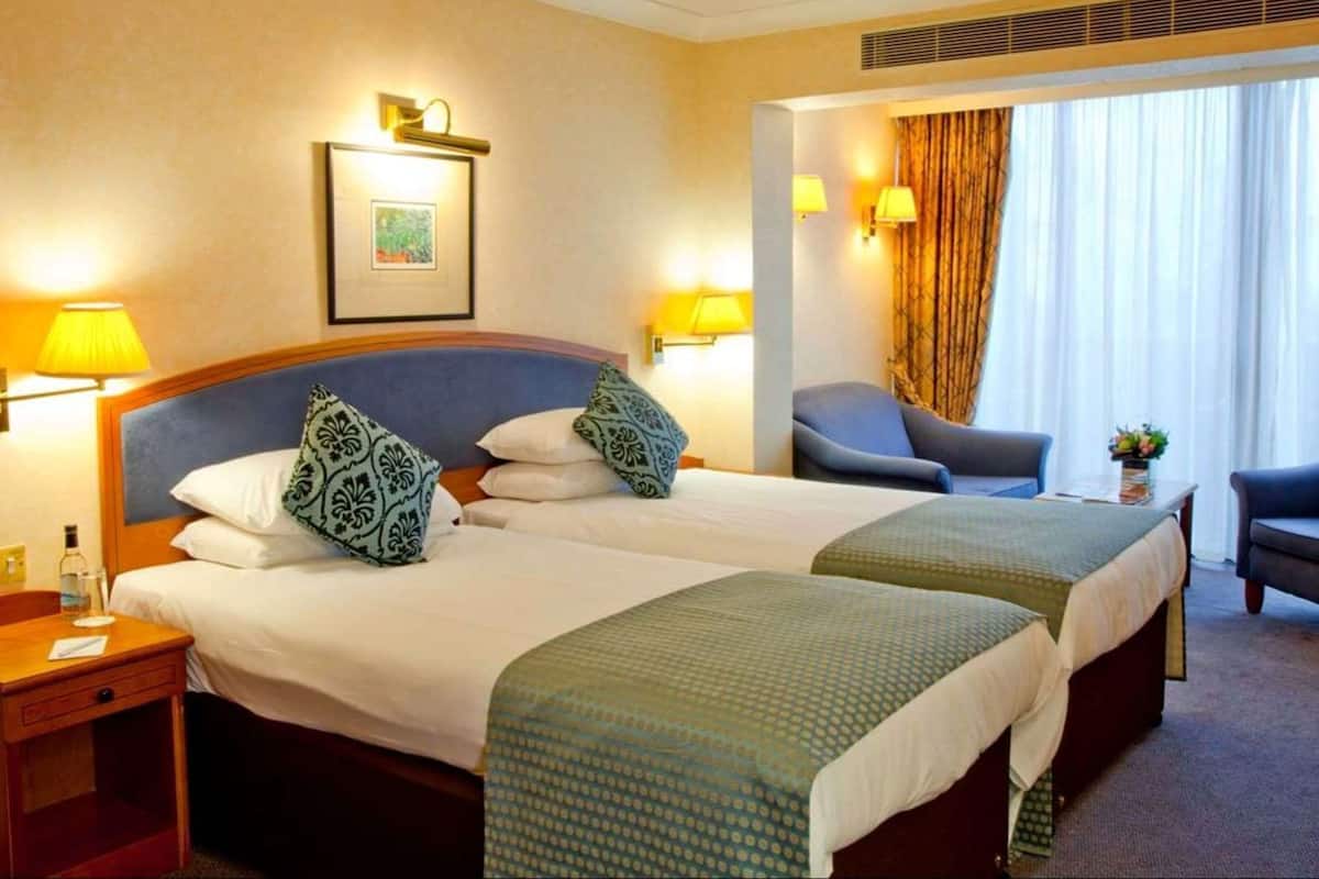 Habitación de hotel con dos camas, una silla, una mesa y un ventanal con cortinas.