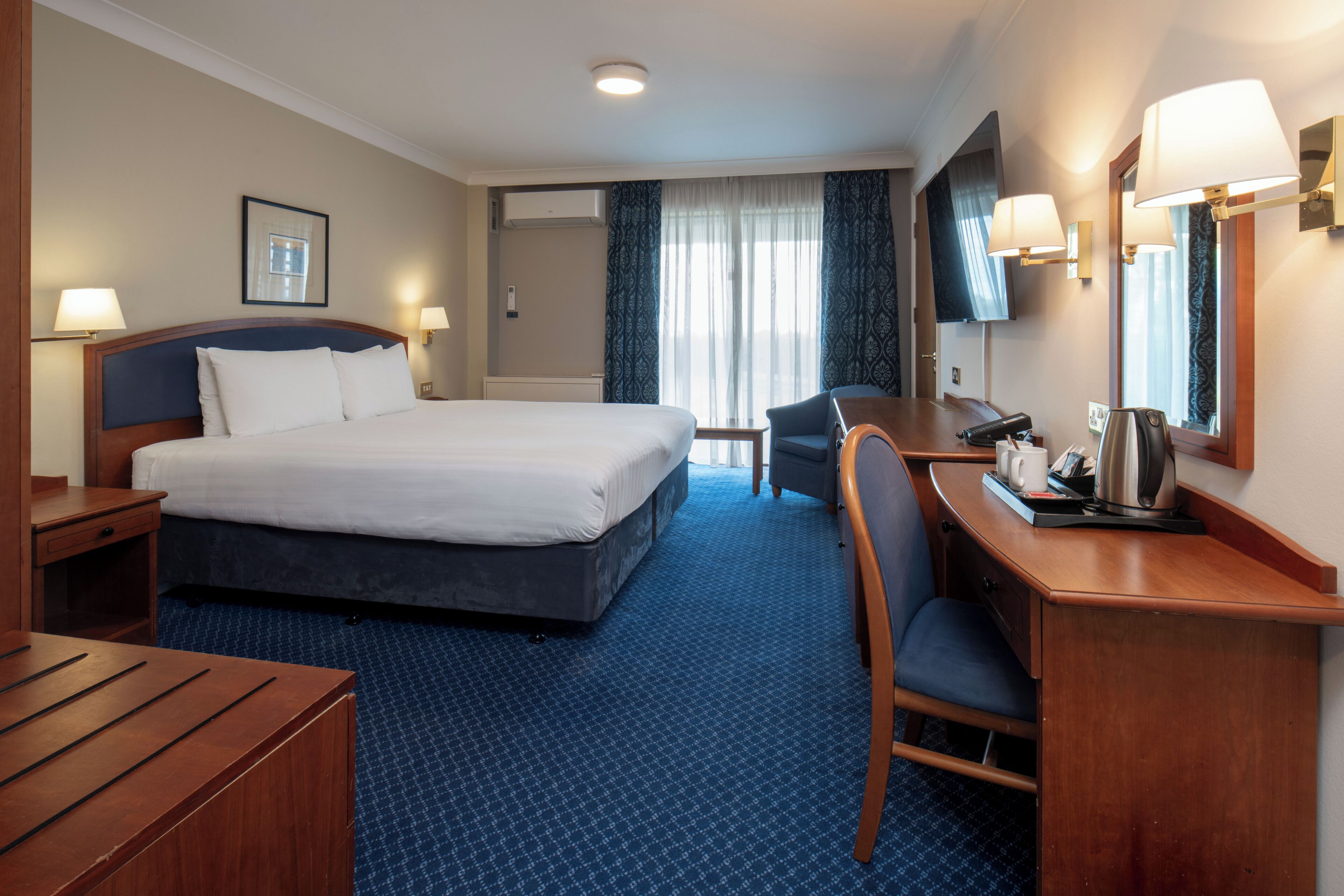 A hotel room with a large bed, a desk, a chair, a flat-screen TV, and a window with curtains.