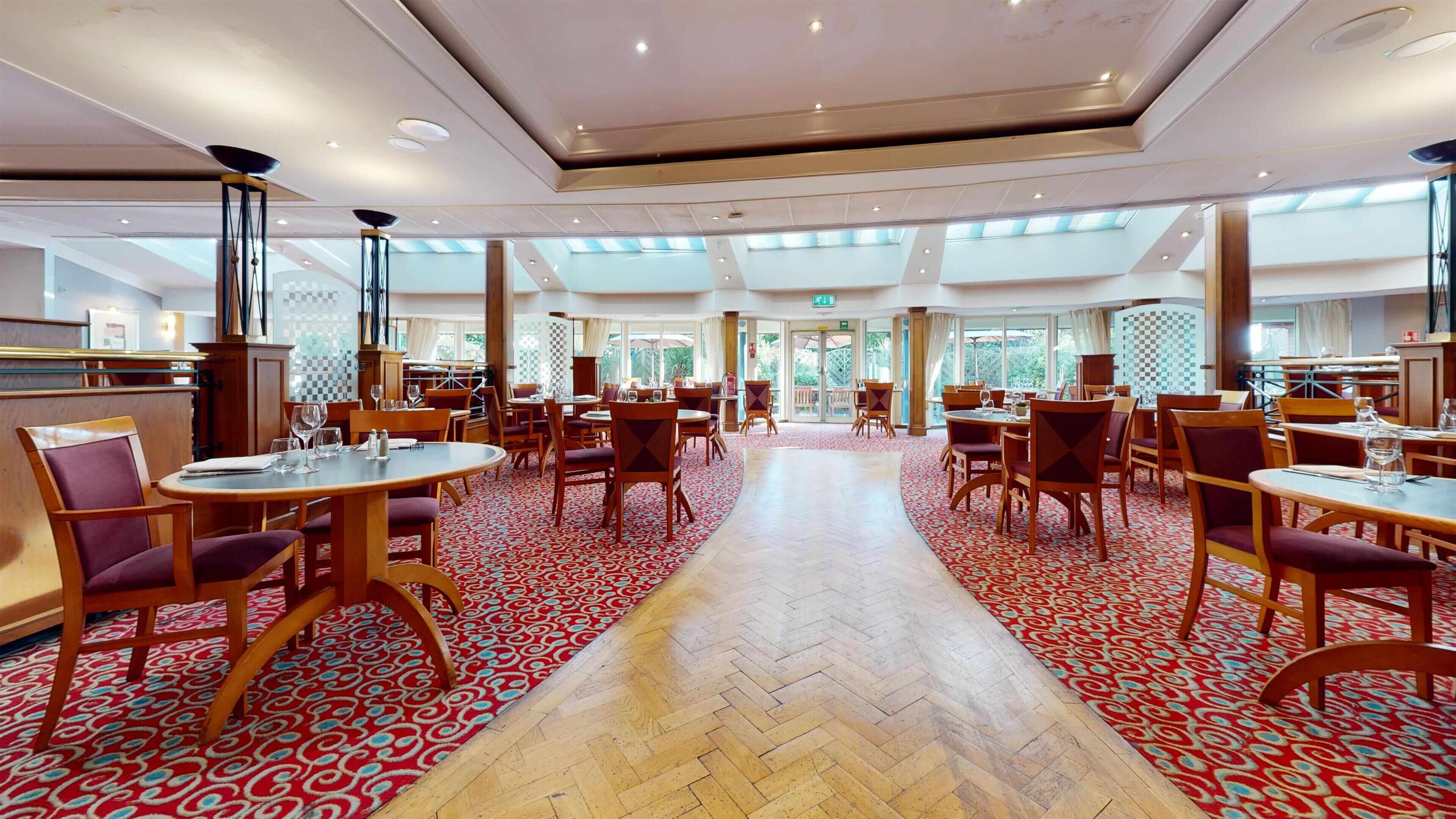 A spacious hotel dining area with multiple tables and chairs, a herringbone patterned floor, and large windows allowing natural light.