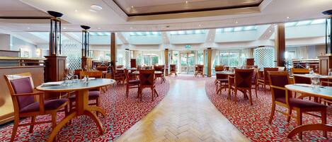 A spacious hotel dining area with multiple tables and chairs, a herringbone patterned floor, and large windows allowing natural light.