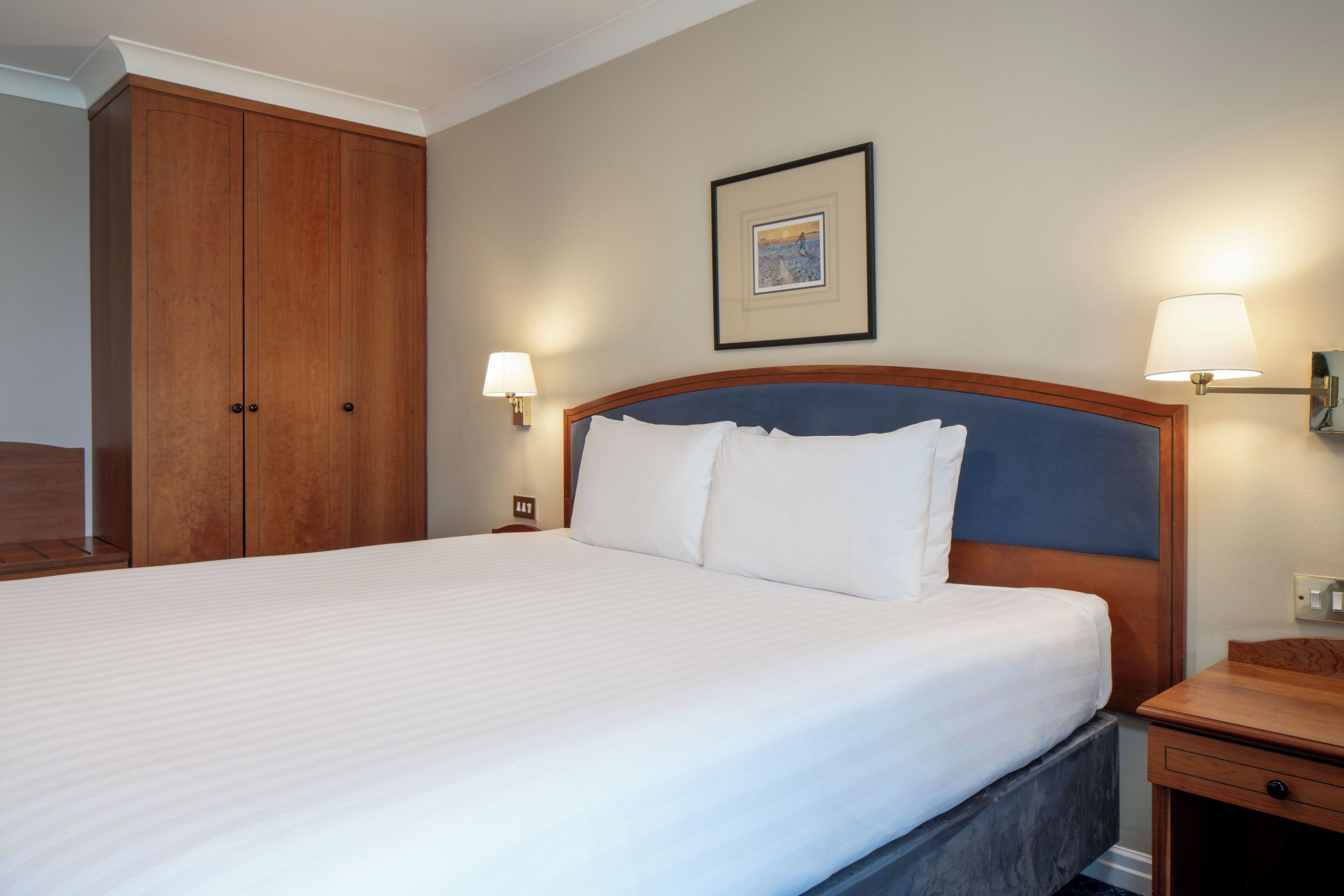 A hotel room with a large bed, wooden wardrobe, two bedside lamps, and a framed picture on the wall.