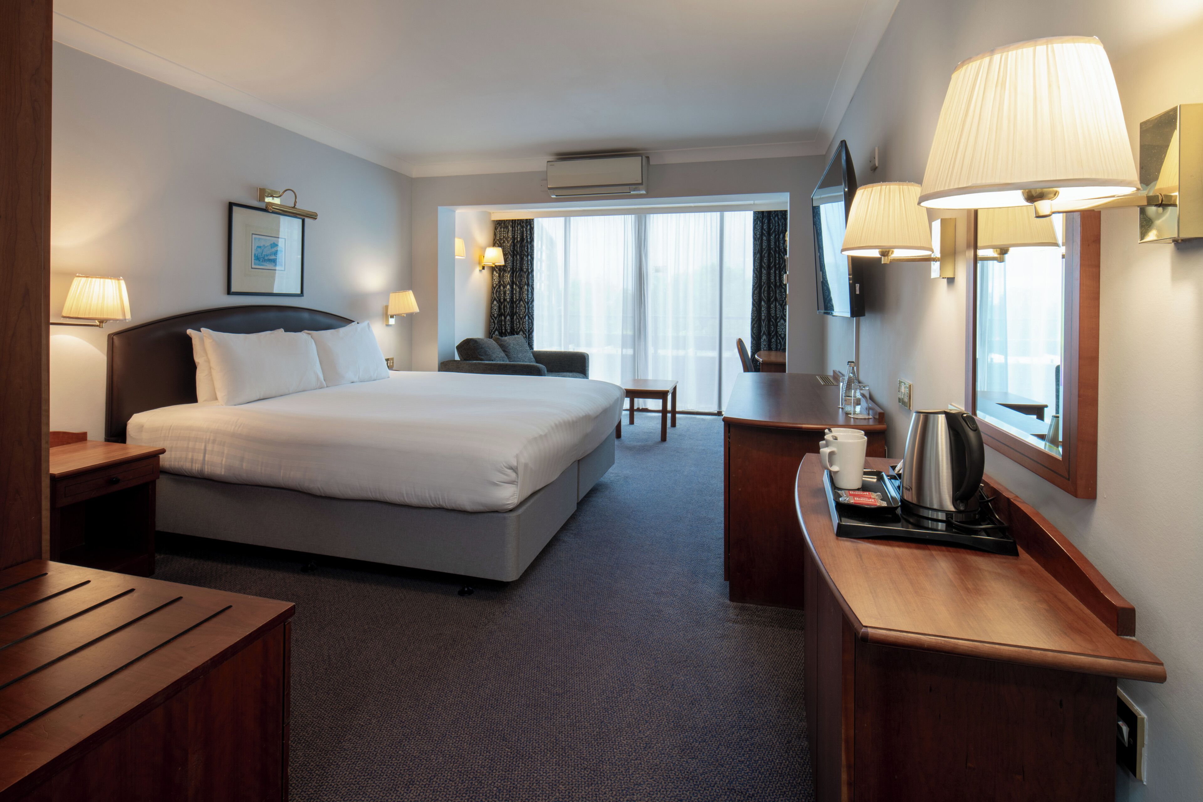 Habitación de hotel con cama, escritorio, silla, cafetera y ventana con cortinas.