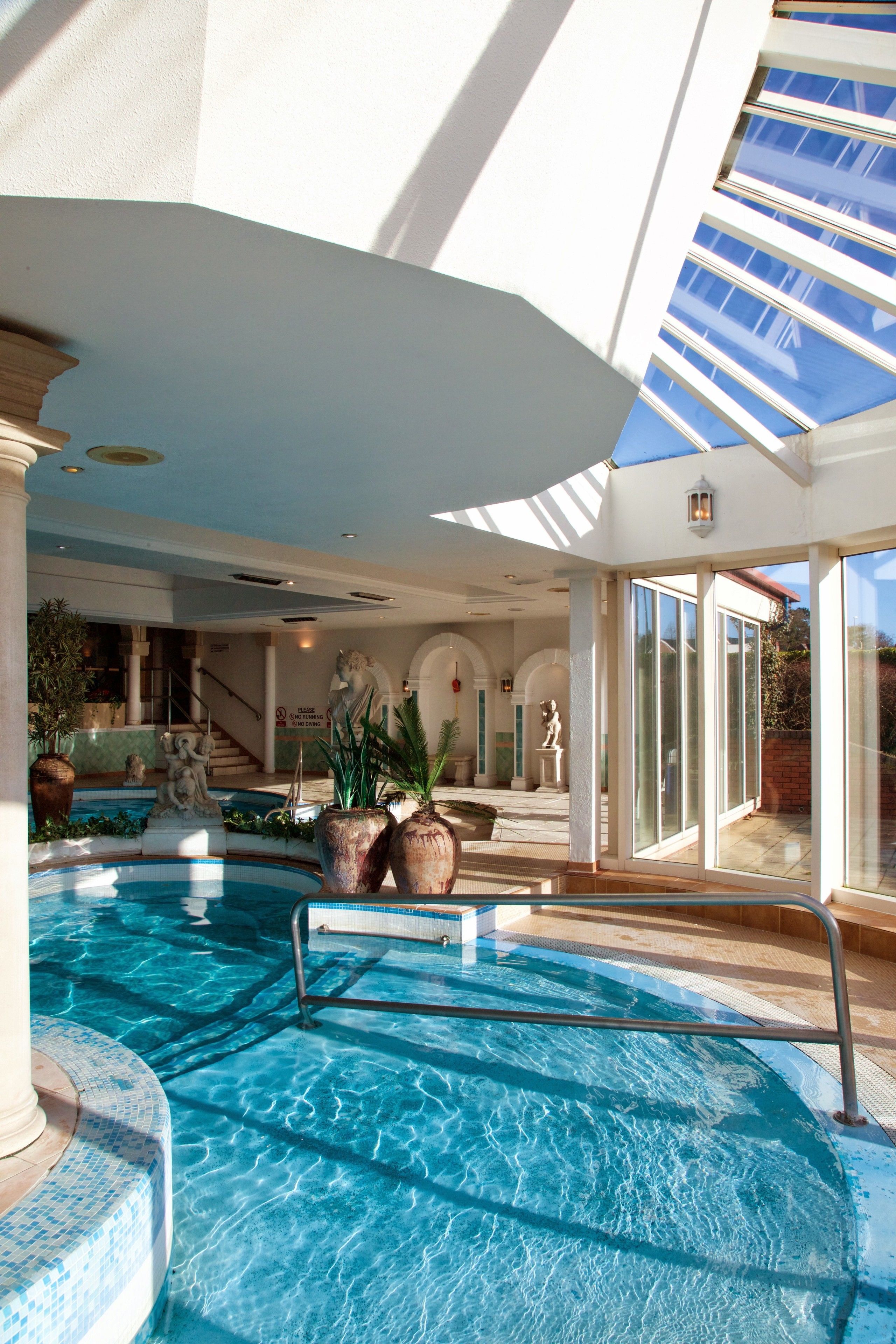 Piscina com teto de vidro, estátuas internas e plantas em vasos.