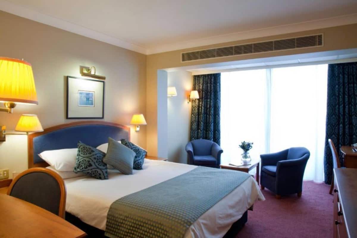 Una habitación de hotel con una cama, dos sillas, un escritorio y un ventanal grande.