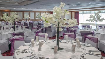 Un salón de banquetes con mesas redondas dispuestas para un evento formal, decorado con flores blancas y cintas moradas.