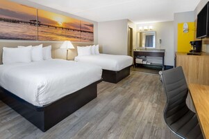 Habitación de hotel con dos camas, un escritorio, una silla, un televisor, un espejo y un baño con lavamanos y toallero.