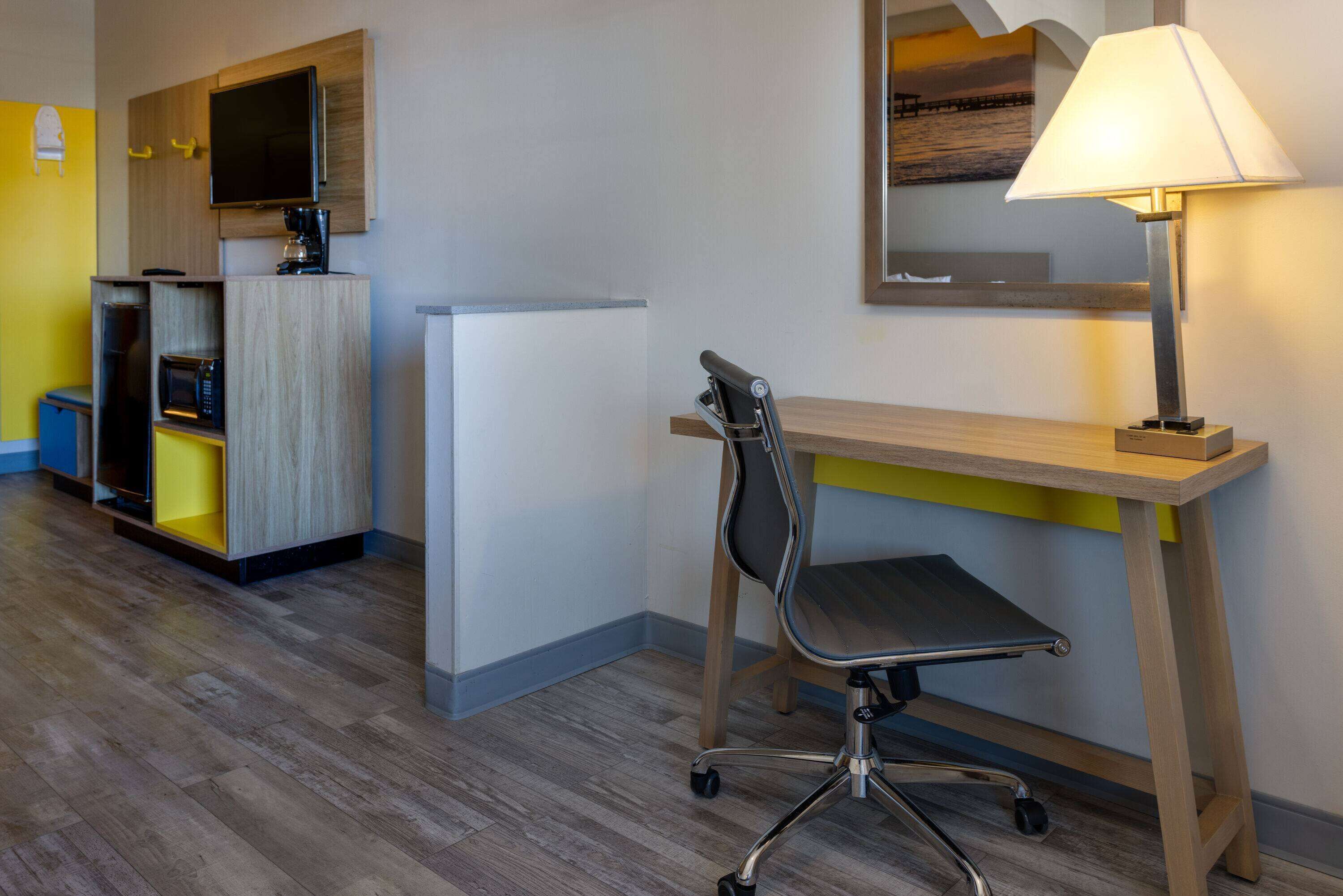 A hotel room with a wooden desk, a chair, a lamp, a television, and built-in kitchen appliances.