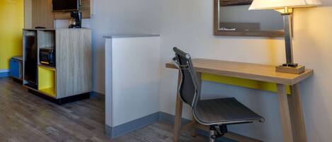 A hotel room with a wooden desk, a chair, a lamp, a television, and built-in kitchen appliances.