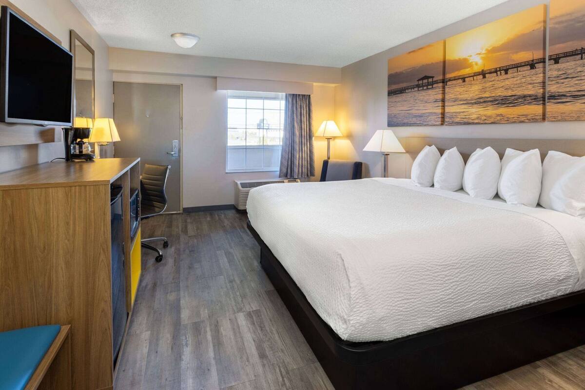 A hotel room with a large bed, a desk with a TV, a window with curtains, and a painting of a sunset over water.
