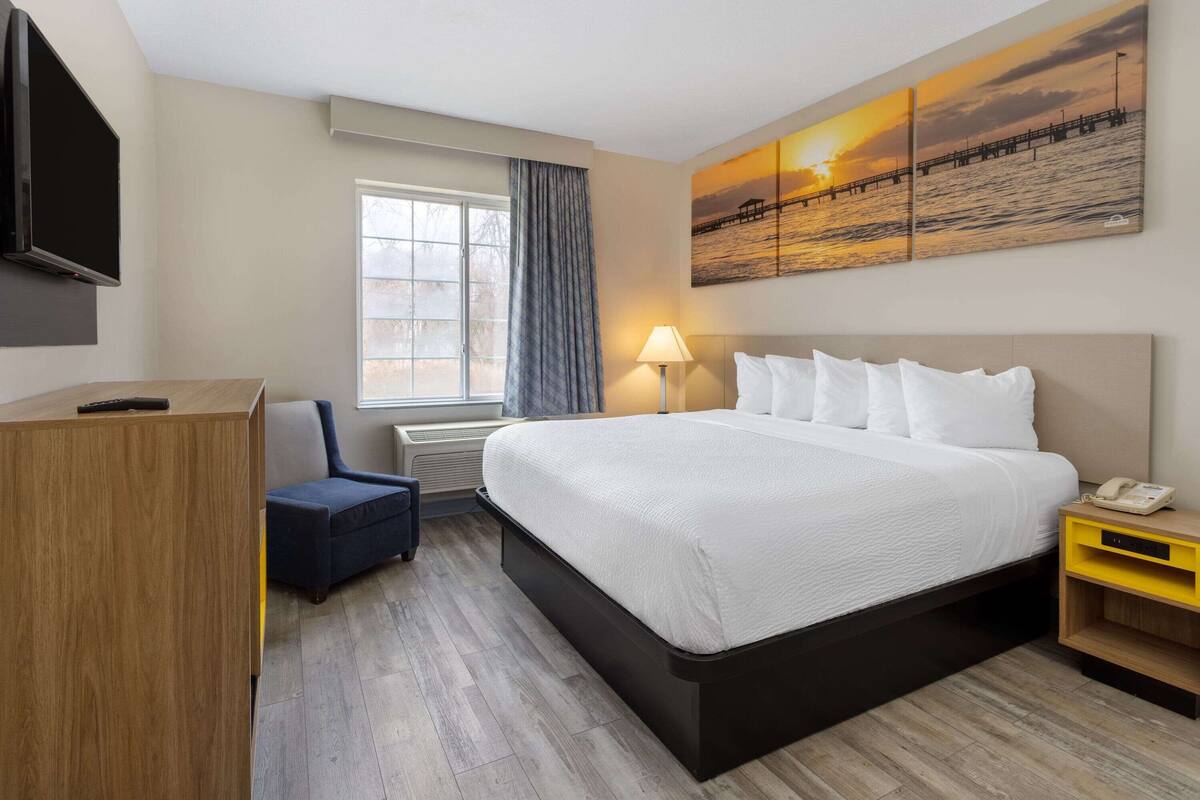 A hotel room with a bed, a desk, a chair, a TV, a lamp, and a painting of a sunset over water.