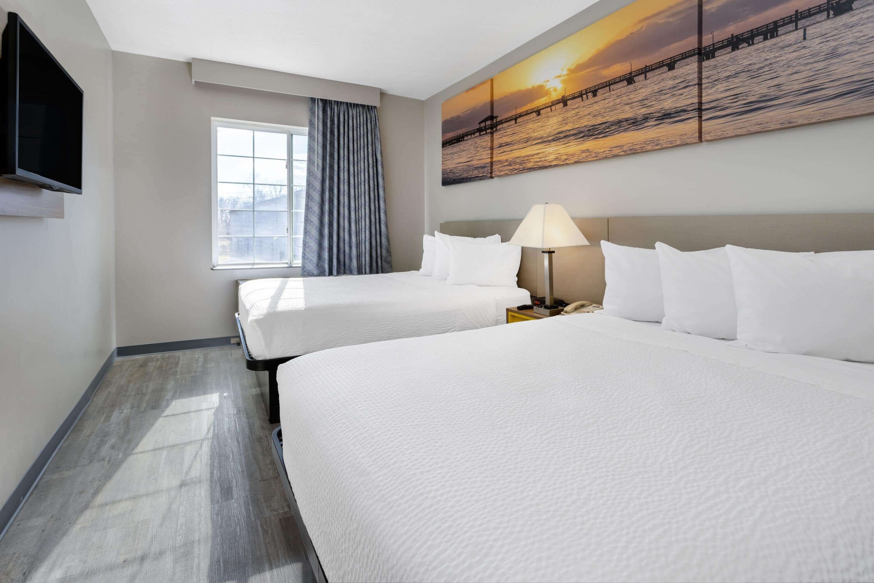 A hotel room with two beds, a TV, a window with curtains, a lamp, and a sunset painting on the wall.