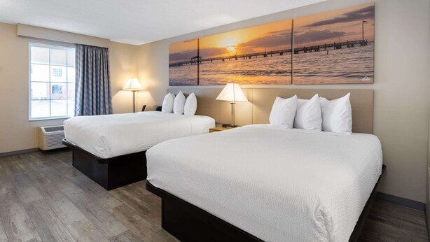 A hotel room with two beds, a large window with curtains, a wall-mounted painting of a sunset over water, and a standing lamp.
