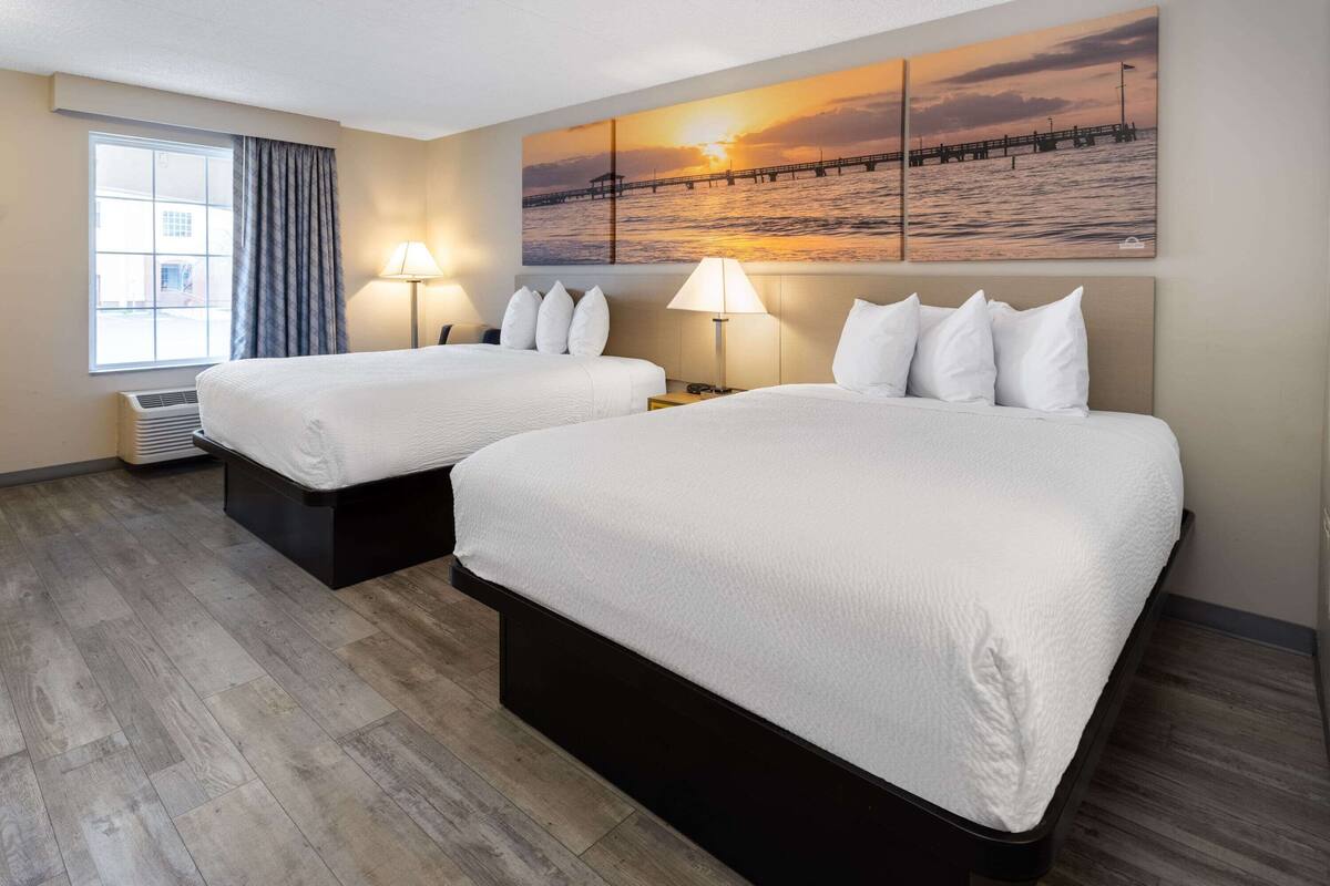 A hotel room with two beds, a large window with curtains, a wall-mounted painting of a sunset over water, and a standing lamp.