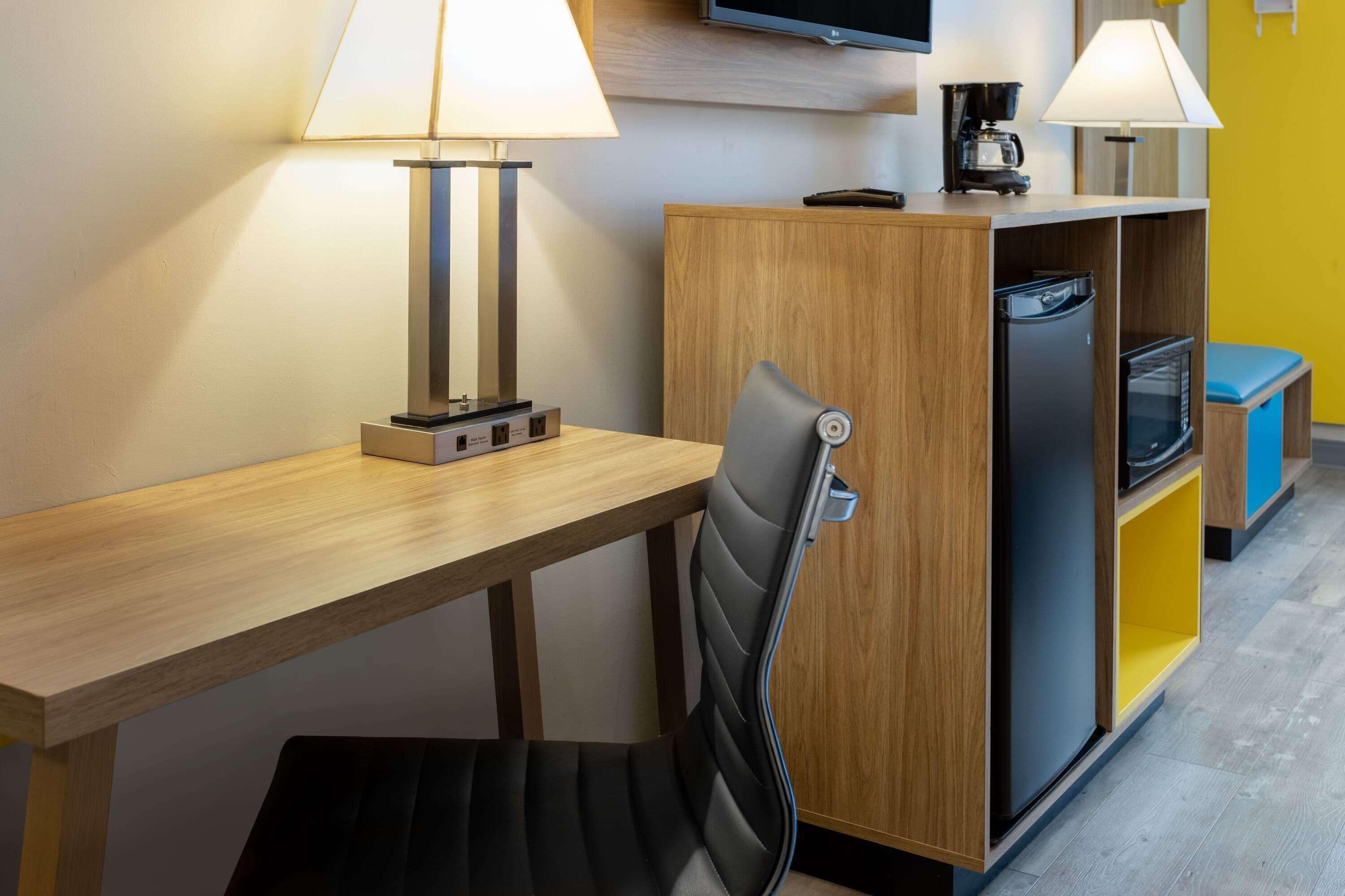 Een kleine hotelkamer met een houten bureau, twee lampen, een stoel, een televisie en een kitchenette met een koelkast en magnetron.