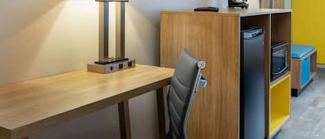 Una habitación de hotel compacta con un escritorio de madera, dos lámparas, una silla, un televisor y una kitchenette con refrigerador y microondas.