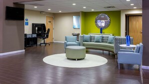 A reception area with a curved sofa, a round ottoman, and a small table with chairs. There is a desk with a computer and a chair, and a television mounted on the wall.
