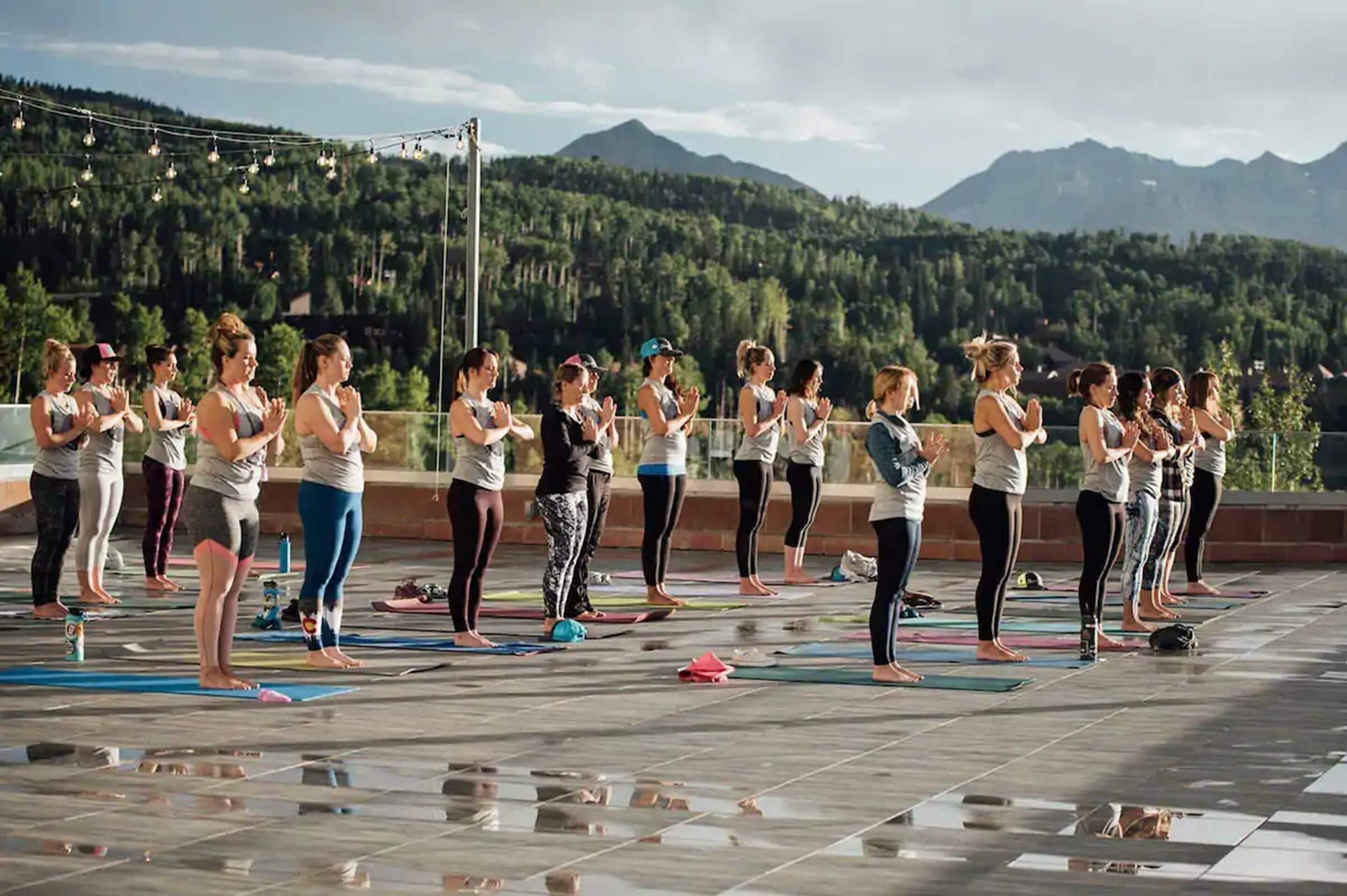 A group of people performing yoga on mats outdoors with a mountainous backdrop.