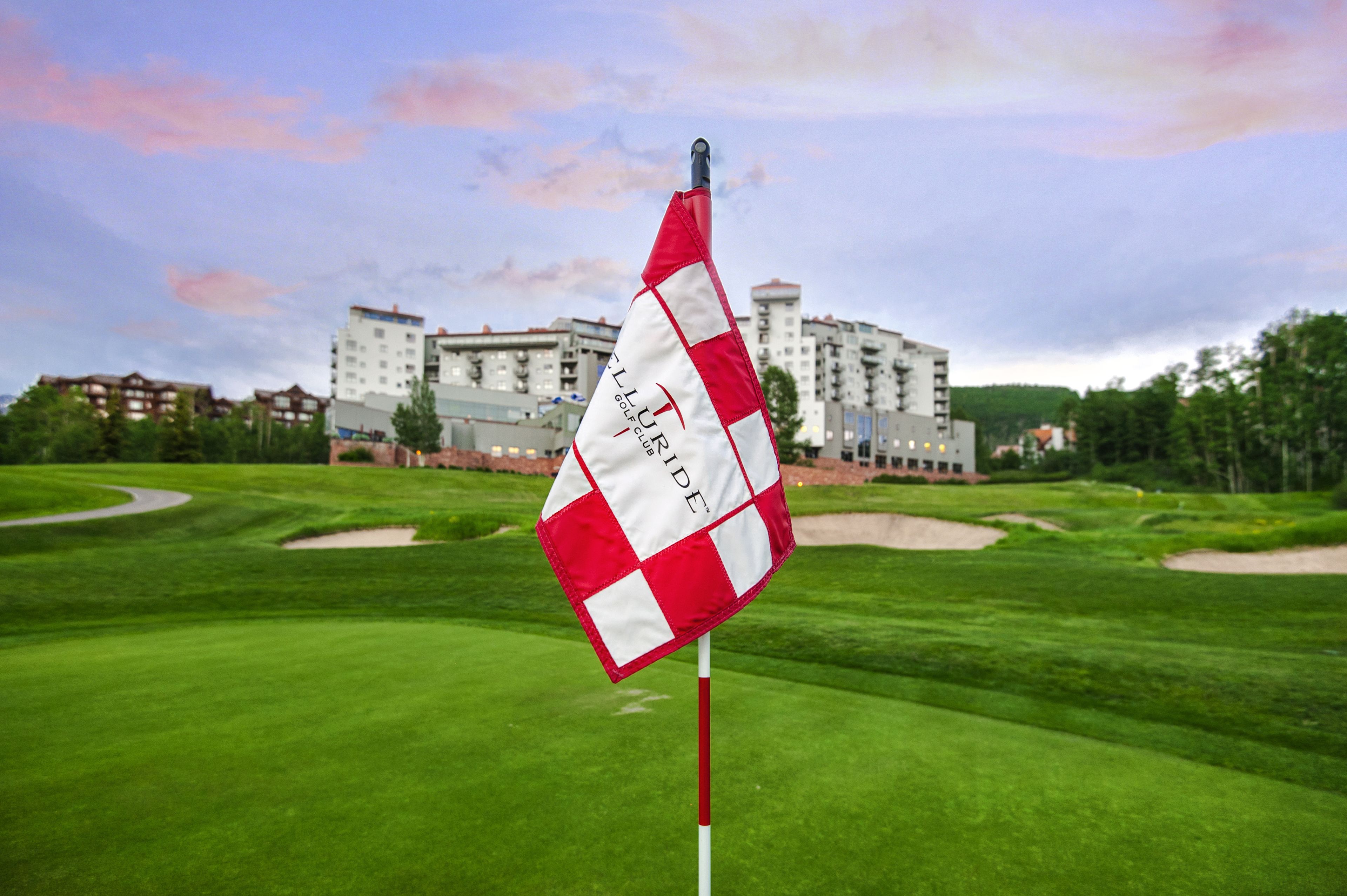 A golf course with a flag on a pole, a building in the background, and a clear sky.