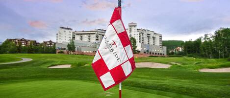 A golf course with a flag on a pole, a building in the background, and a clear sky.