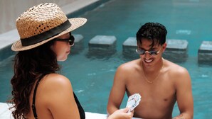 Une personne vêtue d’un bikini noir et d’un chapeau de paille est assise au bord d’une piscine, accompagnée d’une autre personne.
