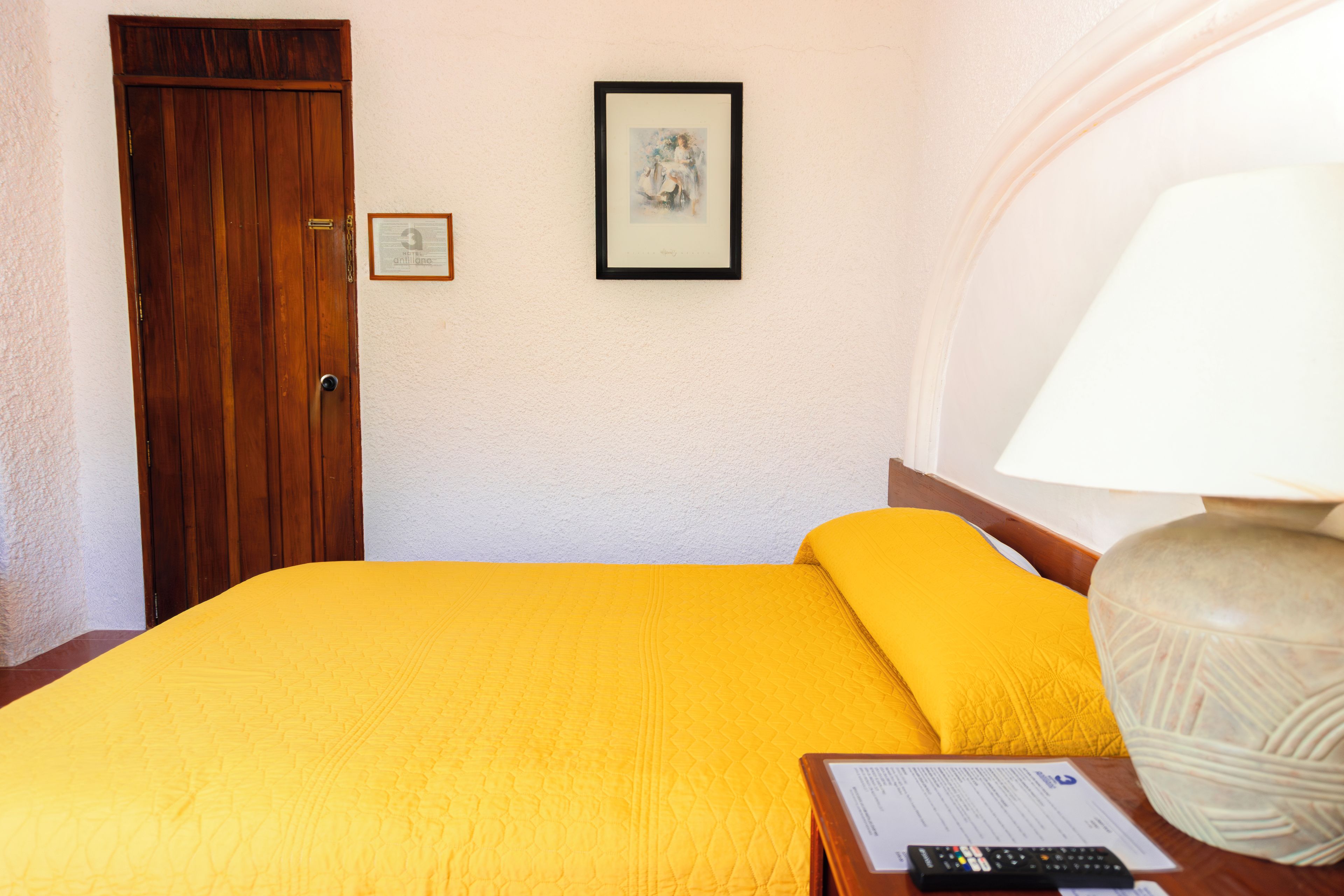 A hotel room with a wooden door, a framed picture on the wall, a yellow bedspread, a bedside lamp, and a remote control on the nightstand.